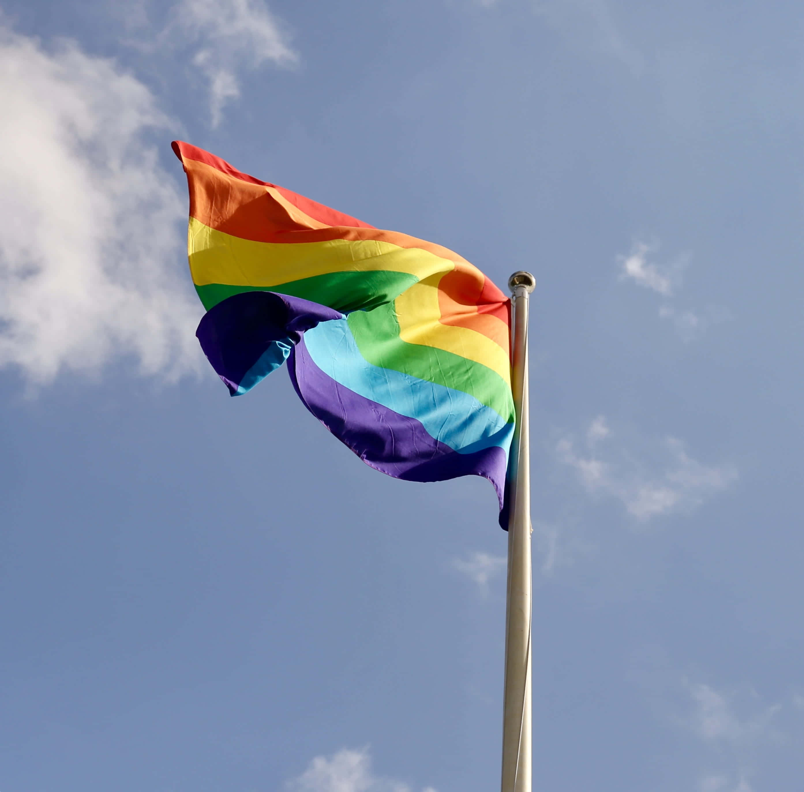 LGBT驕傲旗幟 - 高高飄揚 壁紙 — 一面鮮豔的彩虹旗在藍天白雲下驕傲地飄揚在旗桿上。