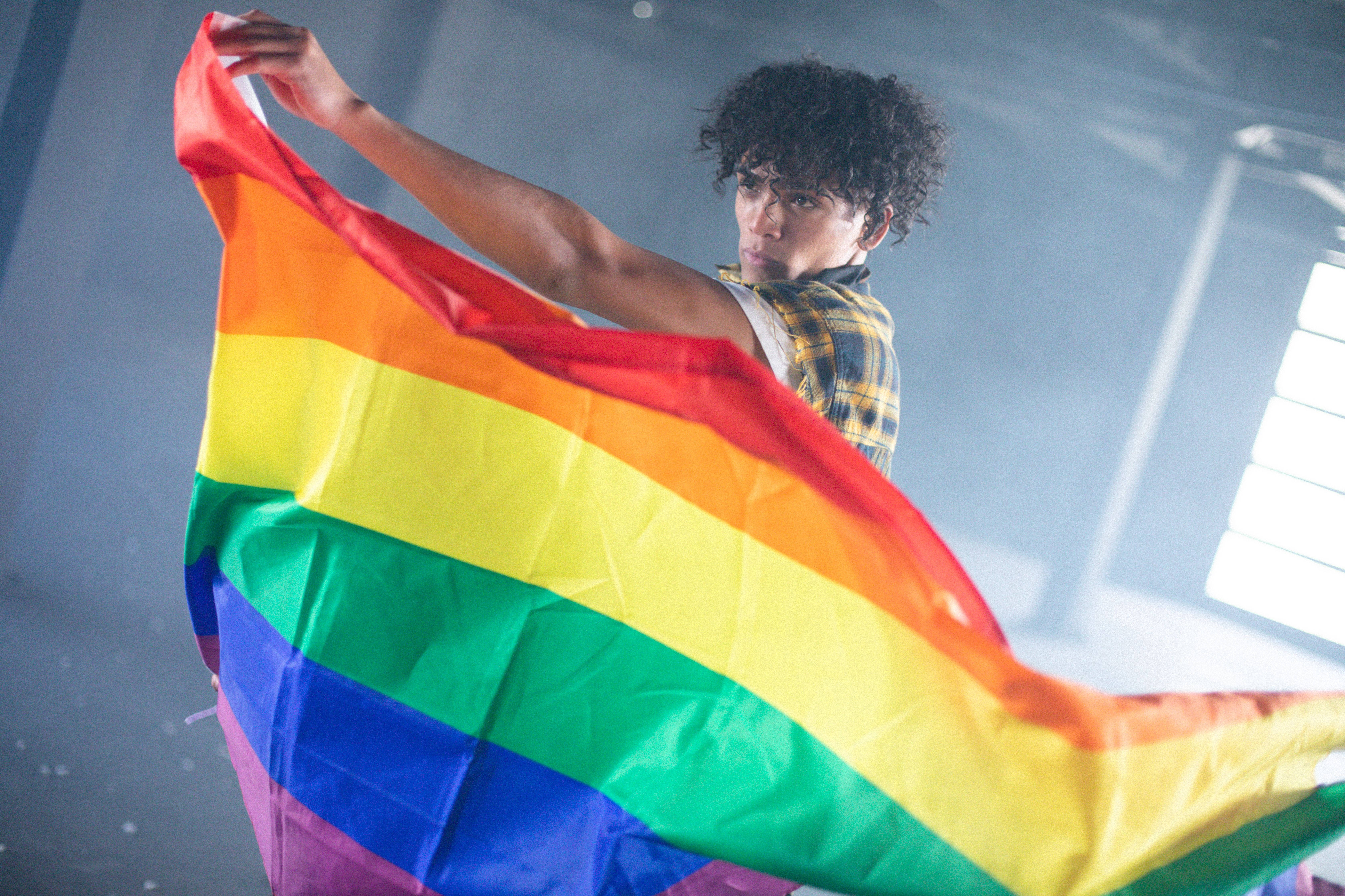 LGBTQ驕傲：多樣性條紋 壁紙 — 一個年輕的捲髮人士在煙霧瀰漫的工業空間中高舉鮮豔的彩虹旗，燈光效果戲劇化。