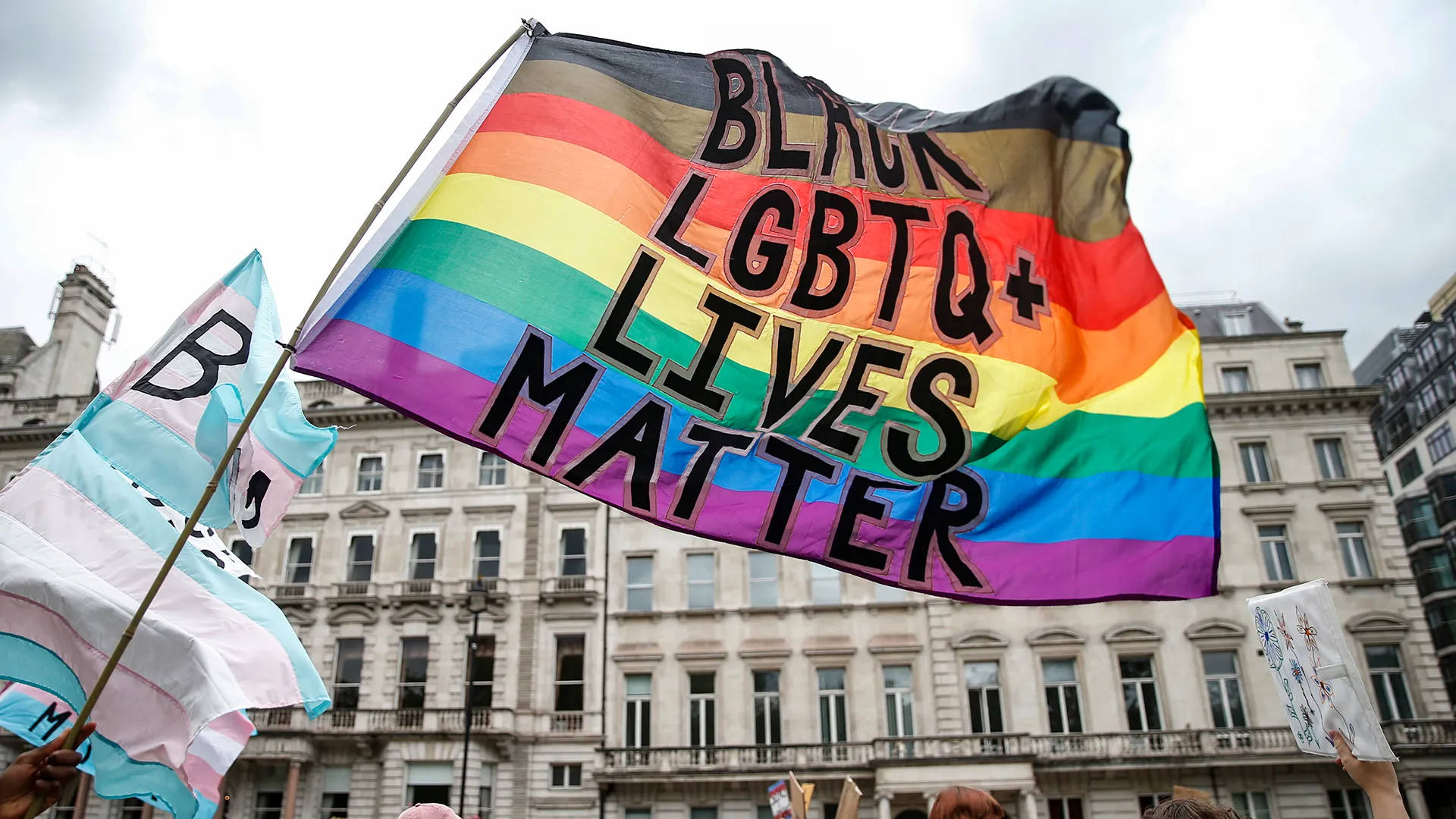 黑人生命至上與LGBTQ+旗幟結合 壁紙 — 一面鮮豔的彩虹旗，上面有粗體黑色文字寫著「BLACK LGBTQ+ LIVES MATTER」，在歷史悠久的城市建築背景下，於抗議活動中自豪地飄揚。