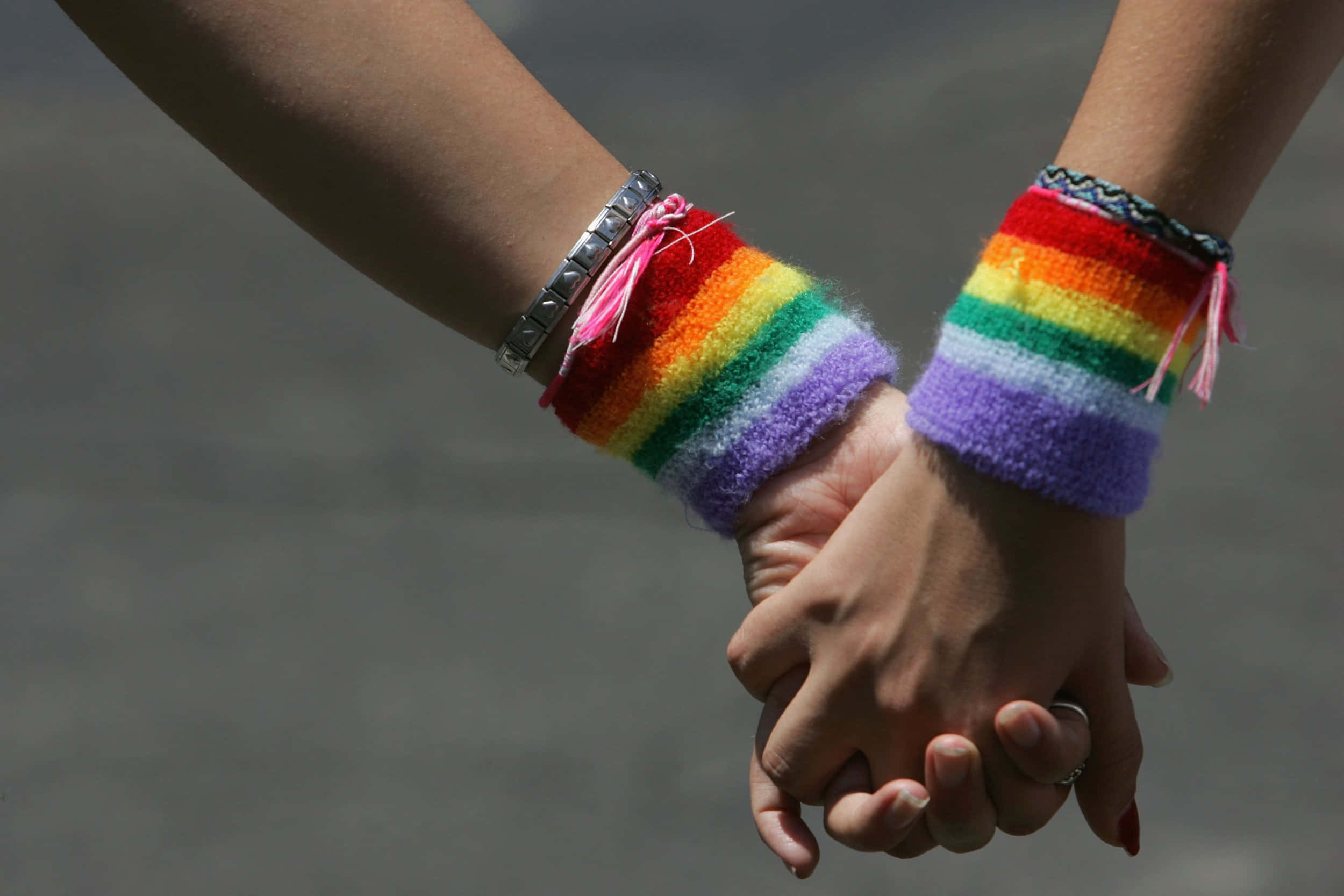 LGBTQ+ 情侶牽手圖片 壁紙 — 兩隻手緊握在一起，戴著色彩繽紛的彩虹編織腕帶，背景是模糊的灰色。