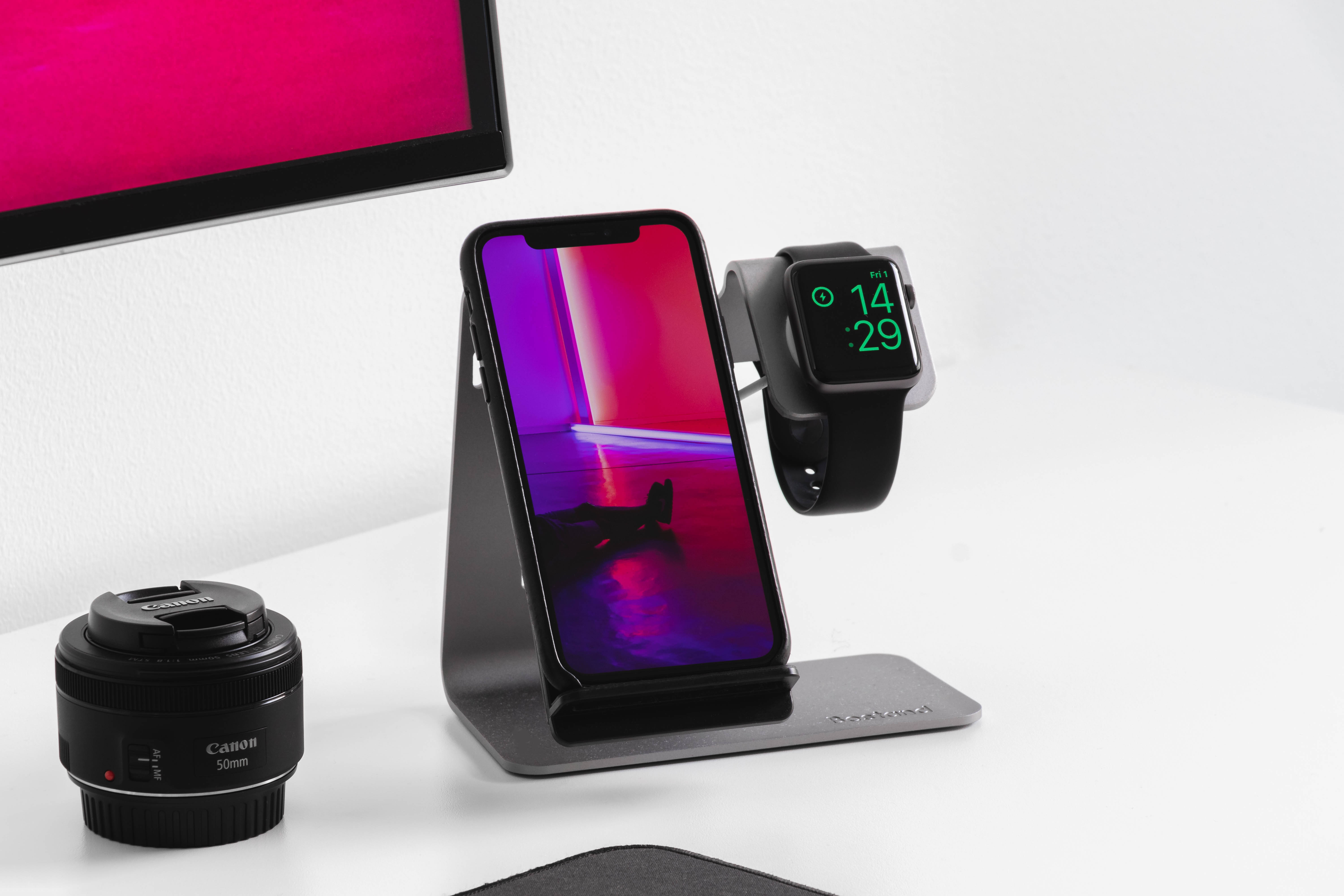 LG手機與Apple Watch 壁紙 — 一部時尚的黑色手機和智能手錶放在充電座上，旁邊是Canon鏡頭和發光的粉紅色顯示器，置於簡約的白色桌子上。