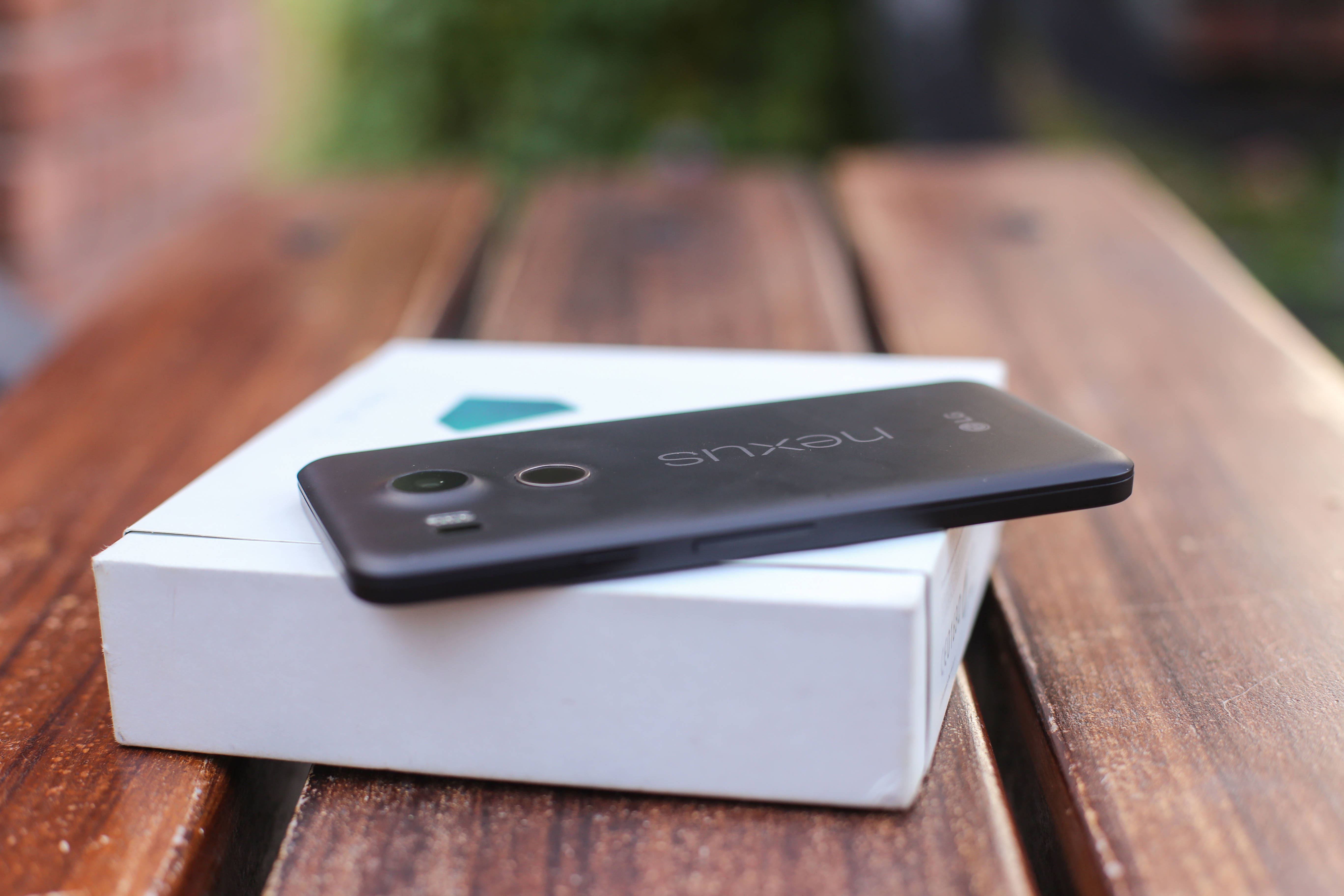 LG手機與Nexus 壁紙 — 一部黑色Nexus智能手機放在其白色盒子上，置於經風化的木桌上，背景有模糊的綠色植物。
