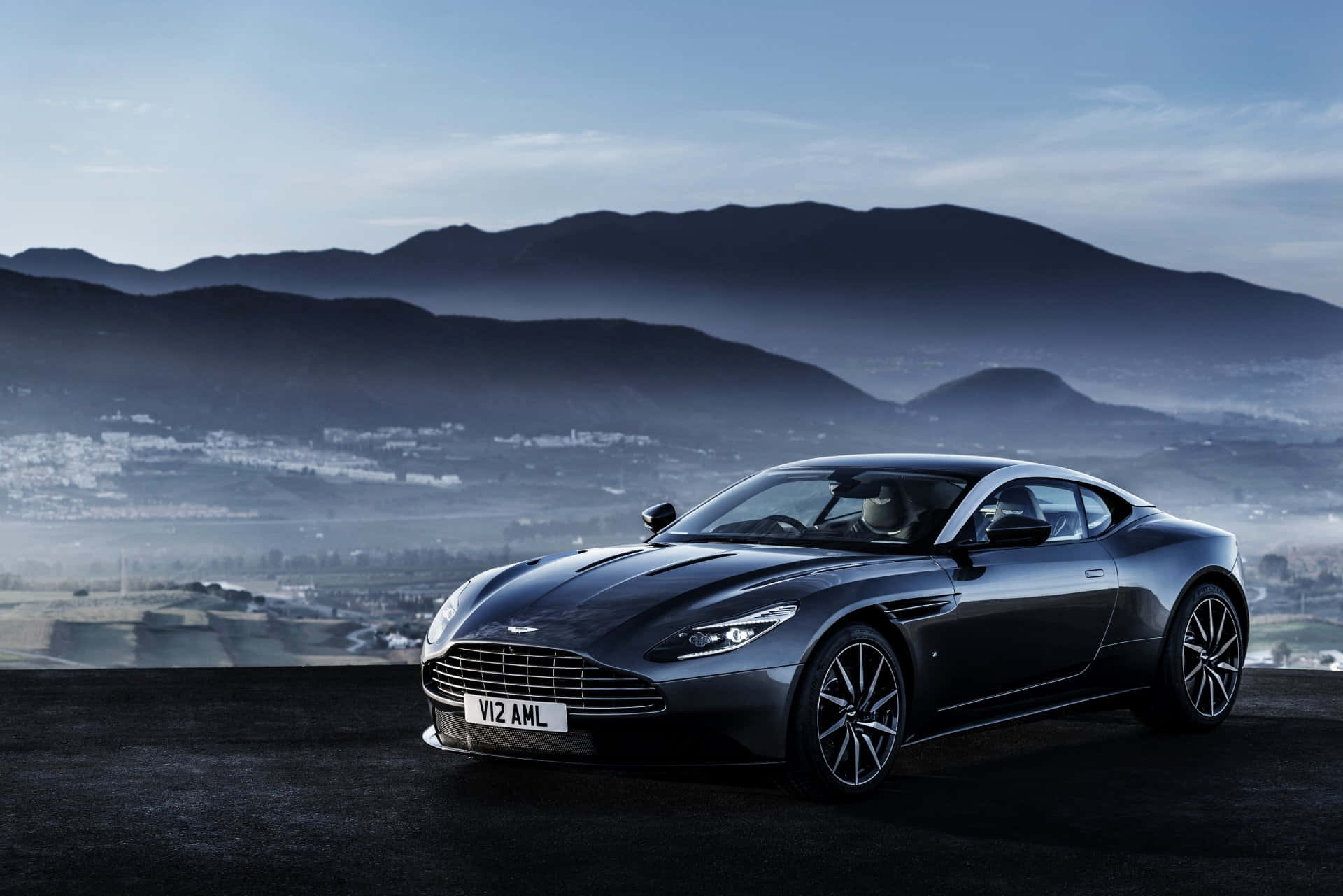 Aston Martin Db11 桌布 壁紙 — 一架時尚的黑色Aston Martin DB11停在懸崖上，俯瞰著黎明時分霧氣繚繞的山脈和山谷小鎮。