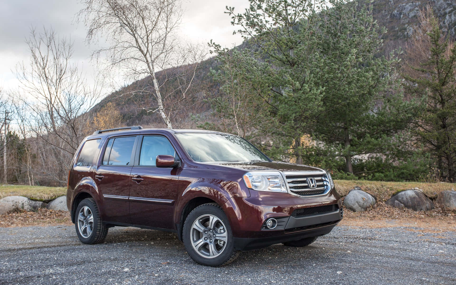 Honda Pilot 桌布 壁紙 — 一輛栗色本田Pilot SUV停泊在碎石上，背景是光禿禿的樹木和岩石山脈，天空多雲。