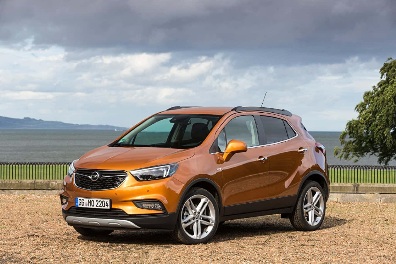 Opel Mokka 桌布 壁紙 — 一架亮橙色Opel Mokka SUV停泊在多雲天空下的海岸圍欄旁的碎石上。