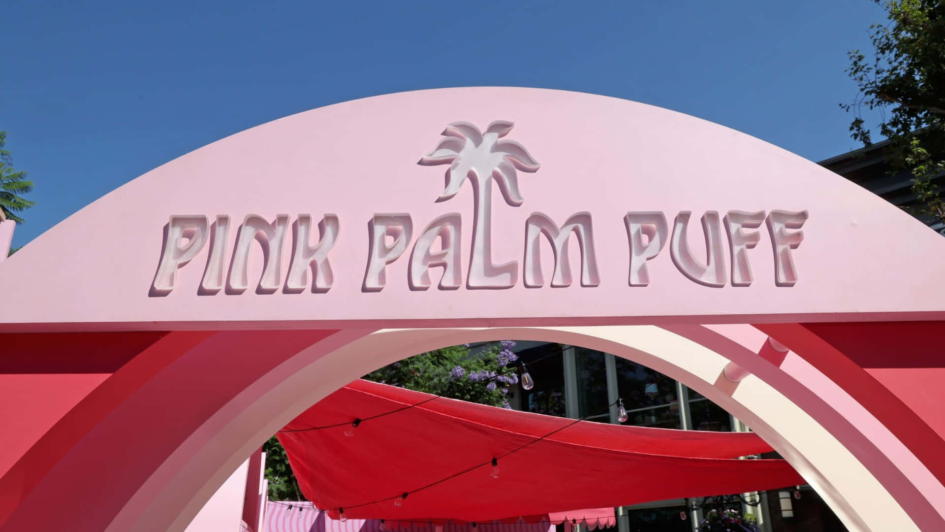  壁紙 — 一個亮粉紅色拱形標誌，上面寫著「PINK PALM PUFF」，並有棕櫚樹標誌，背景是清澈的藍天。