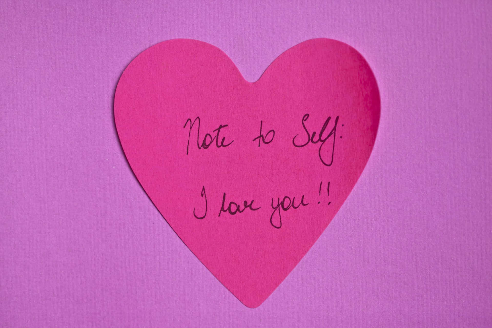  壁紙 — 一個亮粉紅色紙心心，上面有手寫字條寫著「Note to Self: I love you!!」，放在有紋理的紫色背景上。