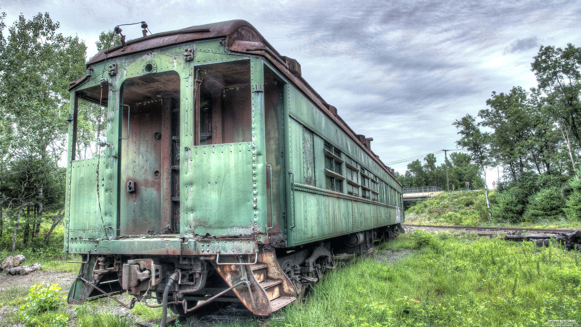  壁紙 — 一輛風化的綠色火車車廂被遺棄在雜草叢生的草地上，天空多雲。