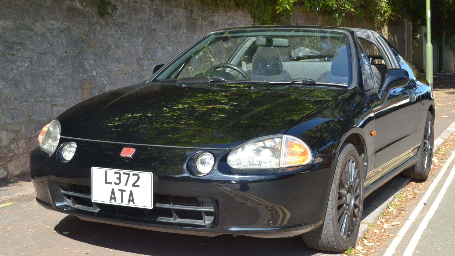 Honda Civic Del Sol 桌布 壁紙 — 一架亮黑色本田Civic Type R停泊在陽光明媚的街道上，旁邊係石牆，上面有綠色樹葉。