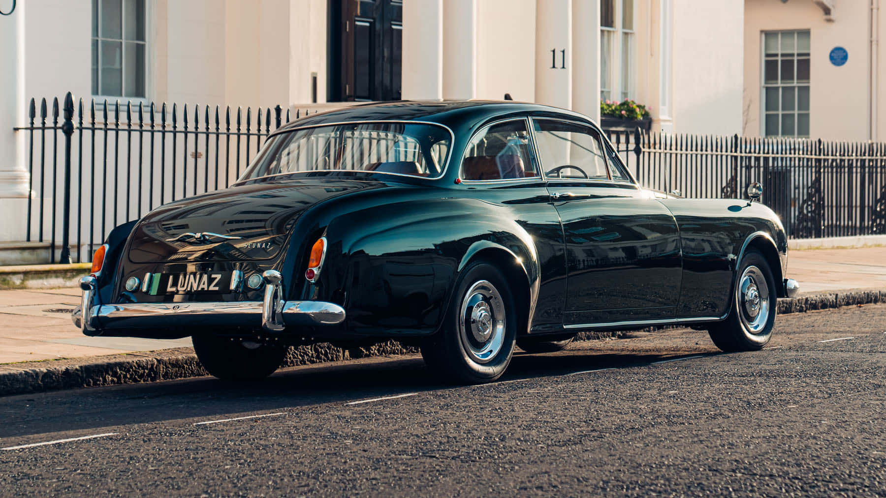 Bentley S2 桌布 壁紙 — 一輛亮黑色復古跑車停泊在陽光明媚的城市街道上，旁邊是白色建築和黑色圍欄。