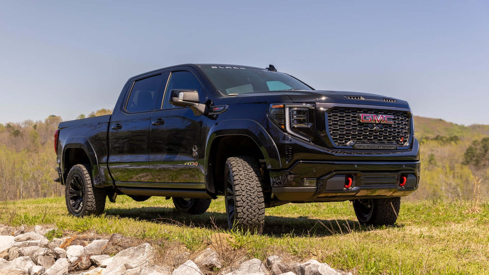 Gmc Sierra 桌布 壁紙 — 一架亮黑色GMC Sierra AT4X pickup truck在晴朗藍天下，堅固地停放在有岩石和遠處樹木的草坡上。