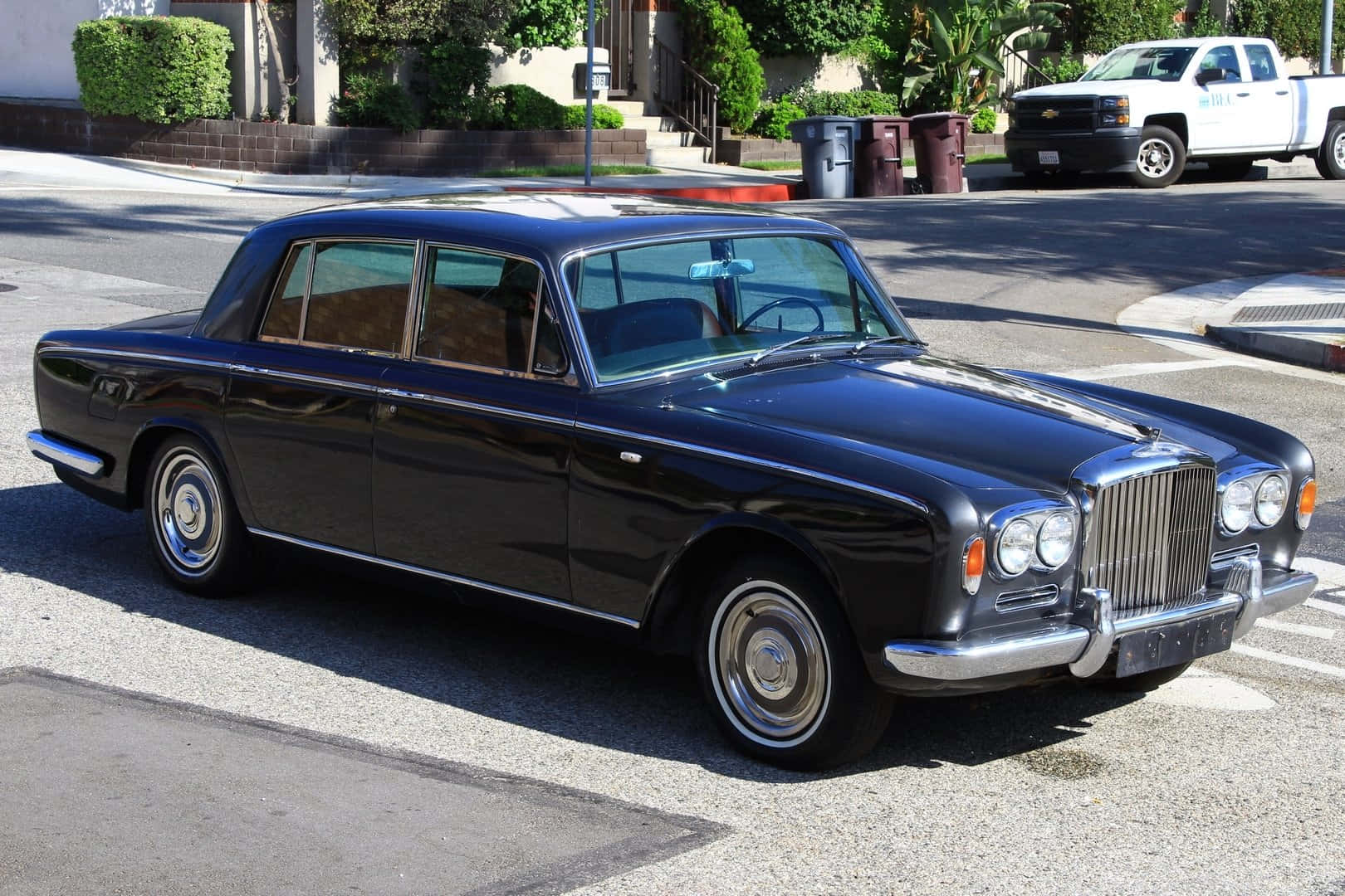 Bentley T1 桌布 壁紙 — 一輛亮黑色 vintage Bentley 房車停泊在陽光普照的住宅街道上，背景有綠化和一輛白色貨車。