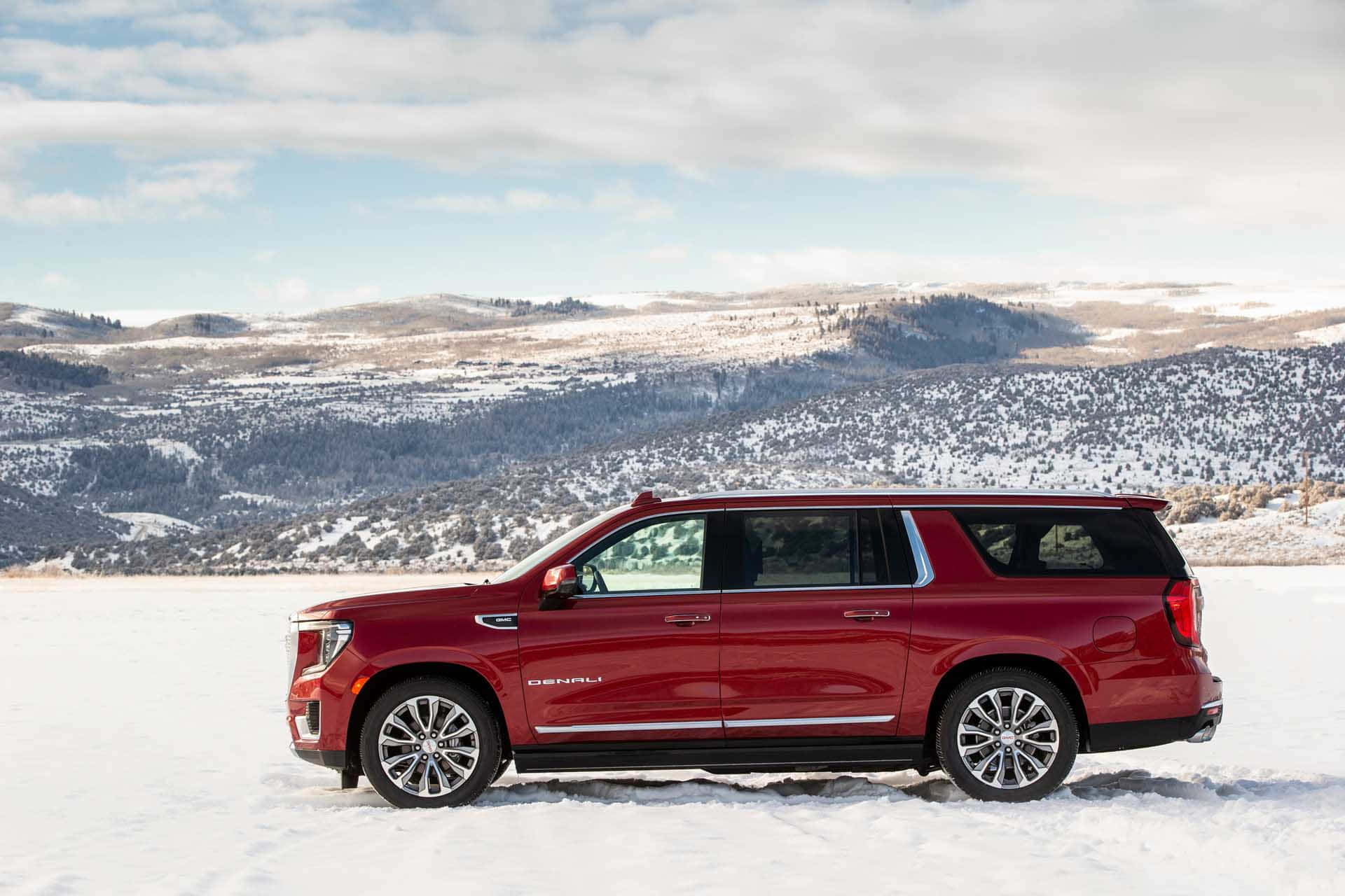 Gmc Yukon 桌布 壁紙 — 一輛亮紅色的Denali SUV停泊在廣闊的雪地上，背景是部分多雲的天空下積雪的山巒。