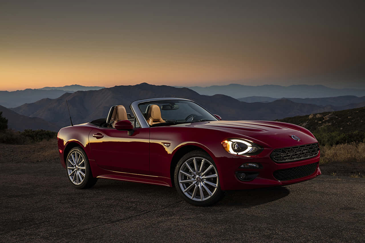 Fiat 124 Spider 桌布 壁紙 — 一架亮紅色開篷跑車停泊在日落時分的山路上，背景是層層疊疊的山脈。