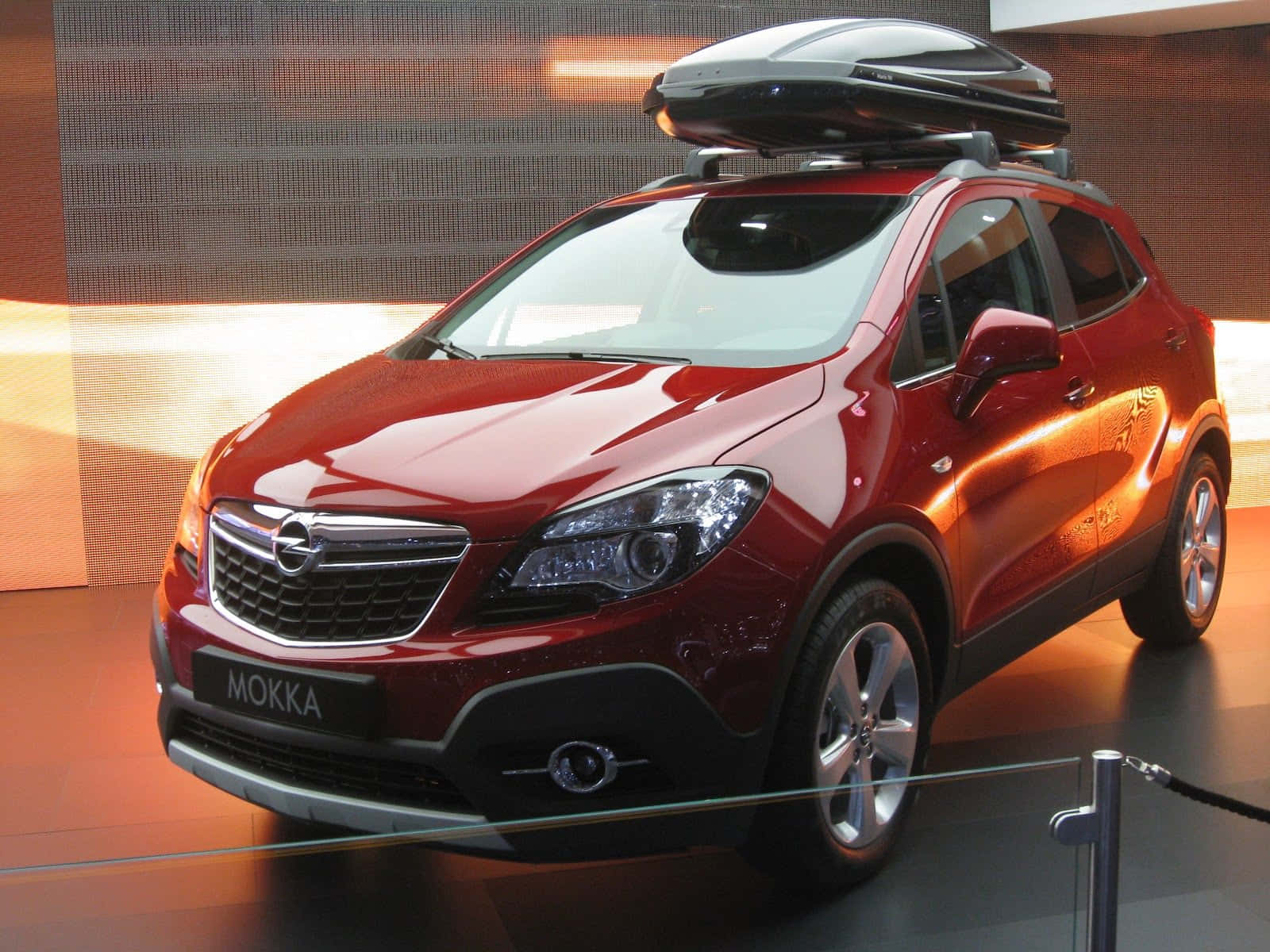 Opel Mokka 桌布 壁紙 — 一輛亮紅色Opel Mokka SUV配備黑色車頂箱，在溫暖的展覽燈光下展示。