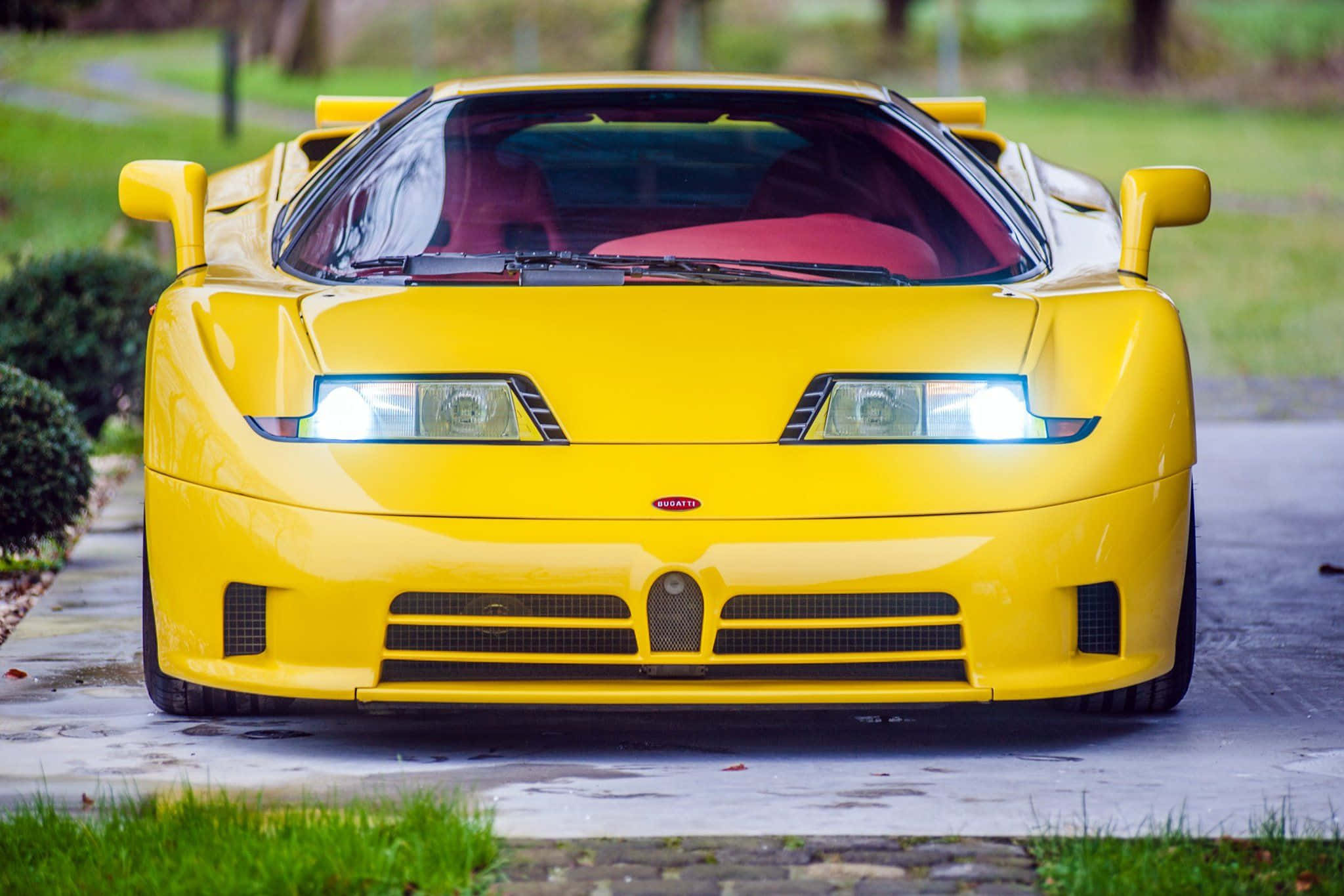 Bugatti Eb110 桌布 壁紙 — 一輛亮黃色的Bugatti EB110超級跑車，亮著車頭燈，停泊在濕漉漉的車道上，周圍環繞著綠草和樹木。