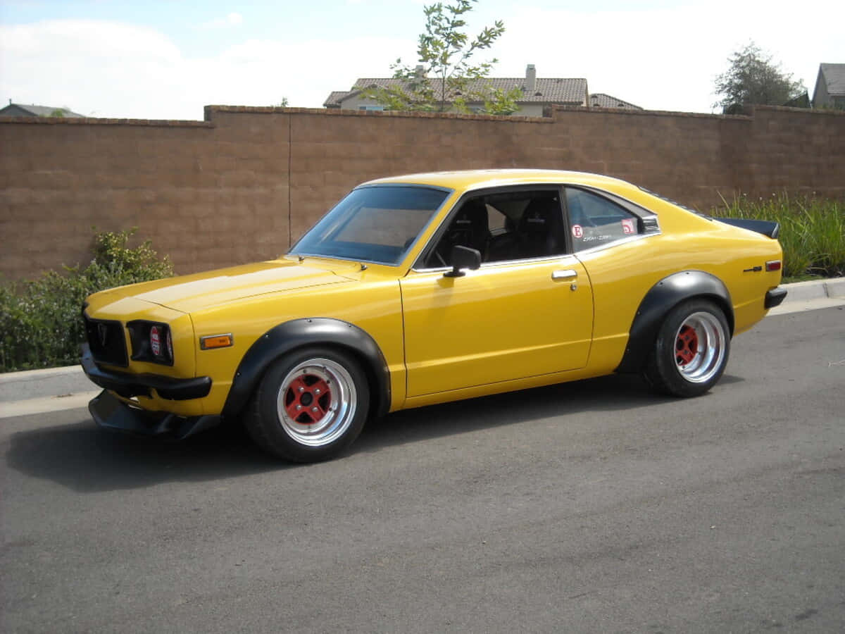 Mazda Rx-3 桌布 壁紙 — 一輛亮黃色的經典日本跑車，紅色輪胎，停在一條磚牆旁邊的街道上。