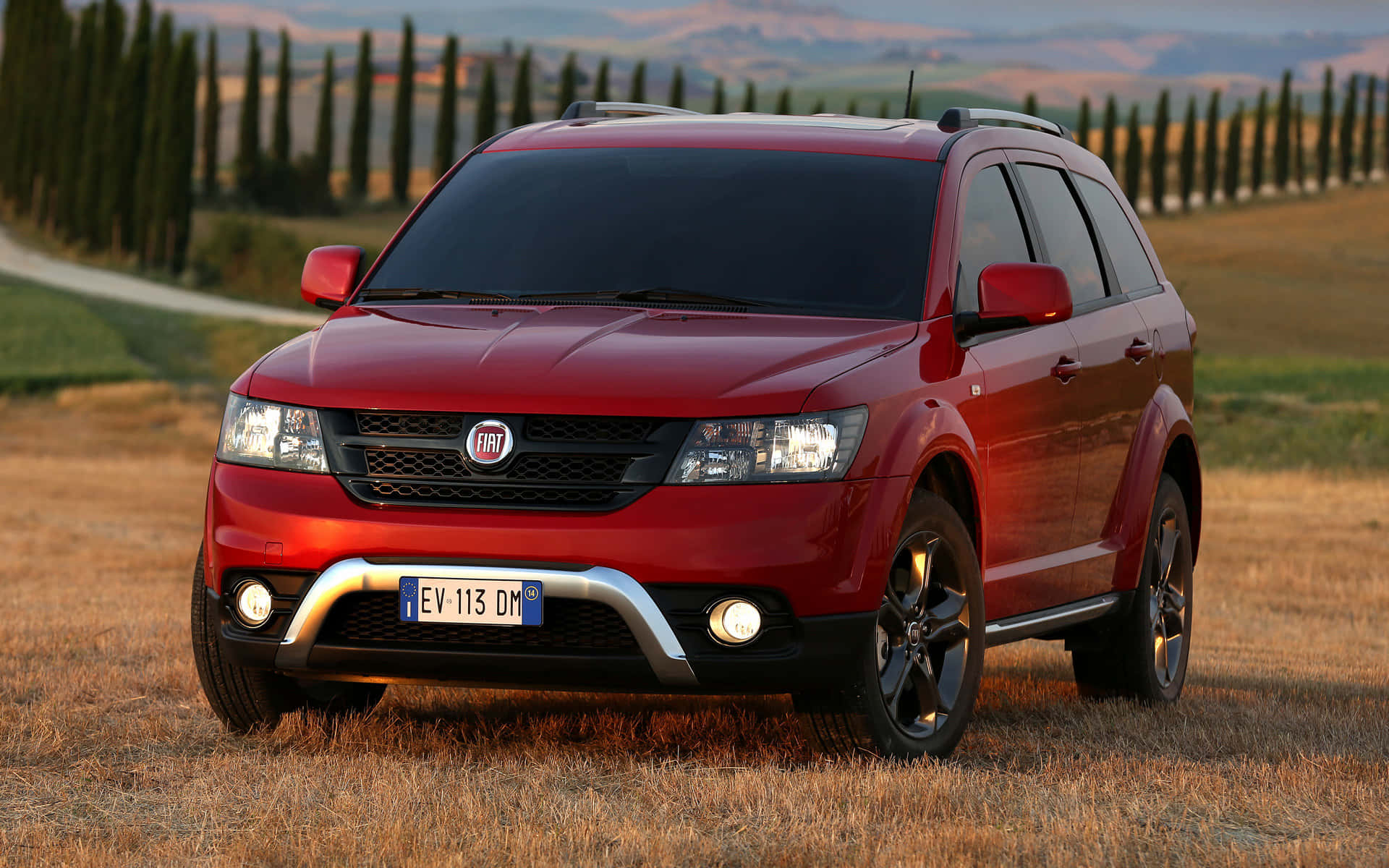  壁紙 — 一架亮紅色嘅Fiat SUV停喺金色田野，柏樹排列喺蜿蜒道路兩旁，背景係溫暖嘅日落天空。