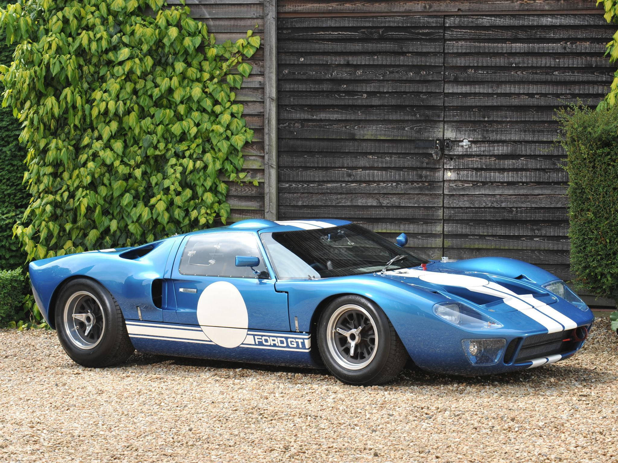 藍色福特GT40賽車，帶有白色條紋，停在碎石地上，靠近木車庫和綠色常春藤。