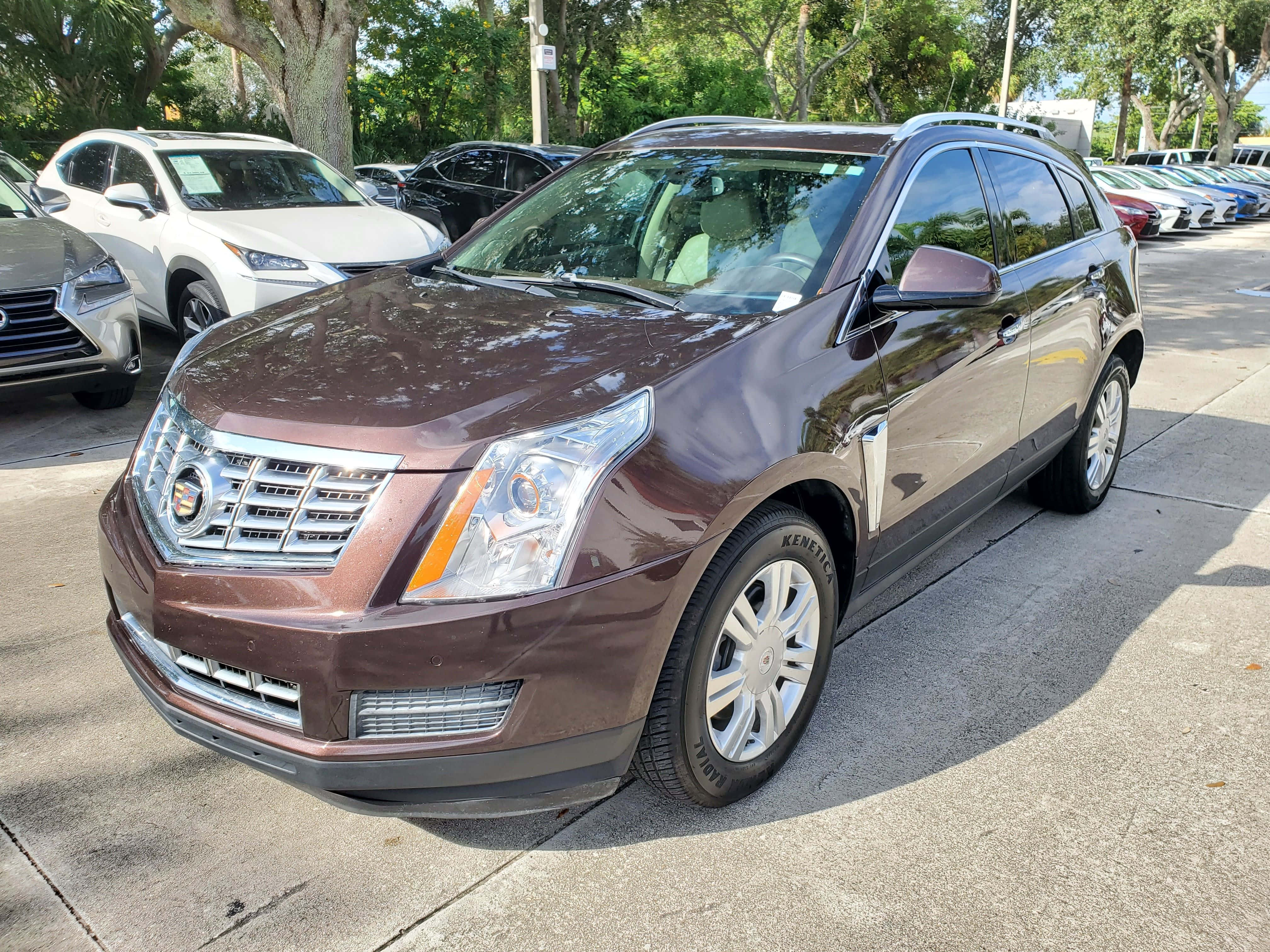 Cadillac Srx 桌布 壁紙 — 一輛亮麗的棕色Cadillac SRX SUV停放在陽光明媚的戶外停車場，背景有樹木和其他車輛。