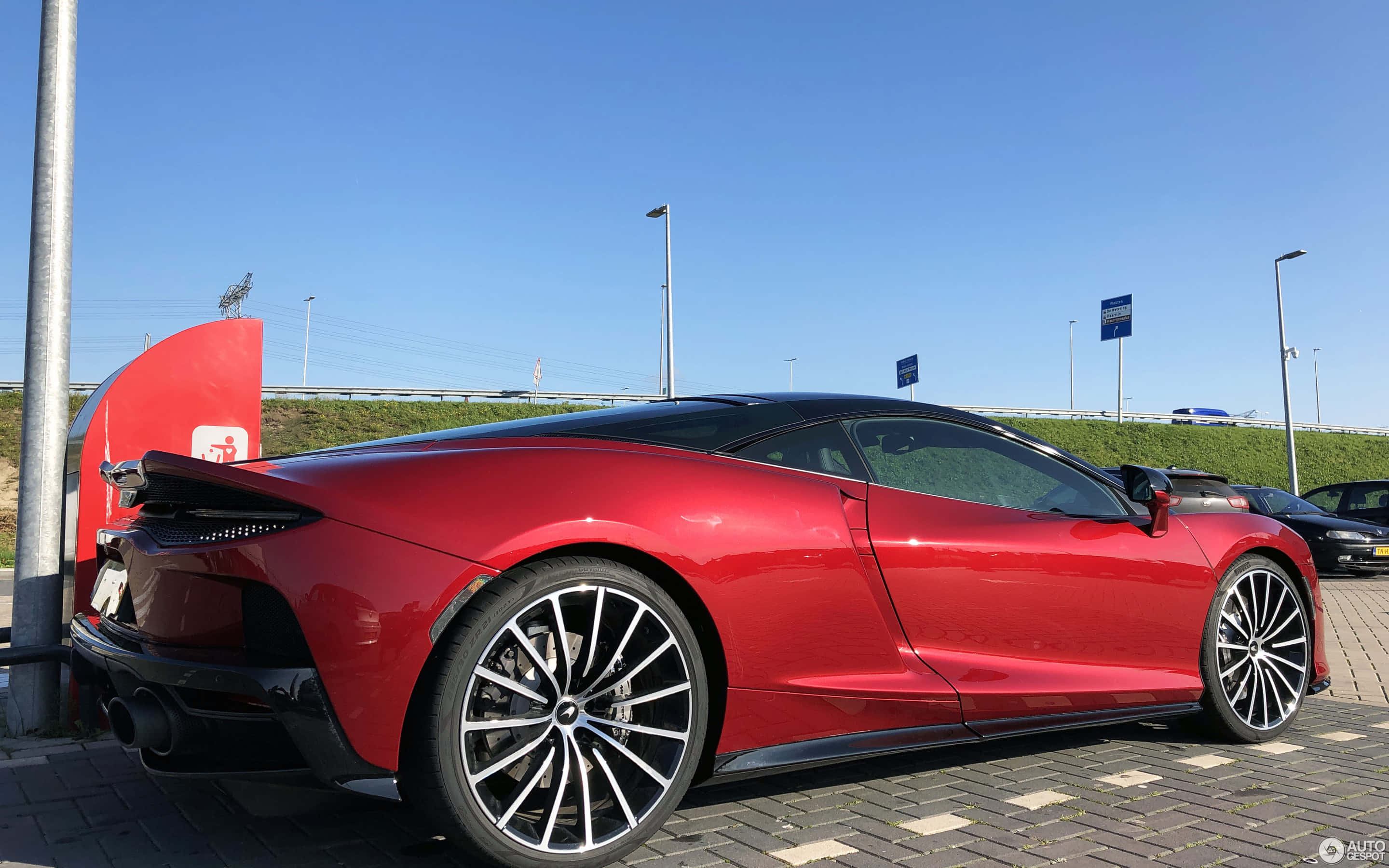 Mclaren Gt 桌布 壁紙 — 一架亮麗的紅色McLaren跑車停泊在鋪砌的停車場上，背景是晴朗的藍天和長滿草的路堤。