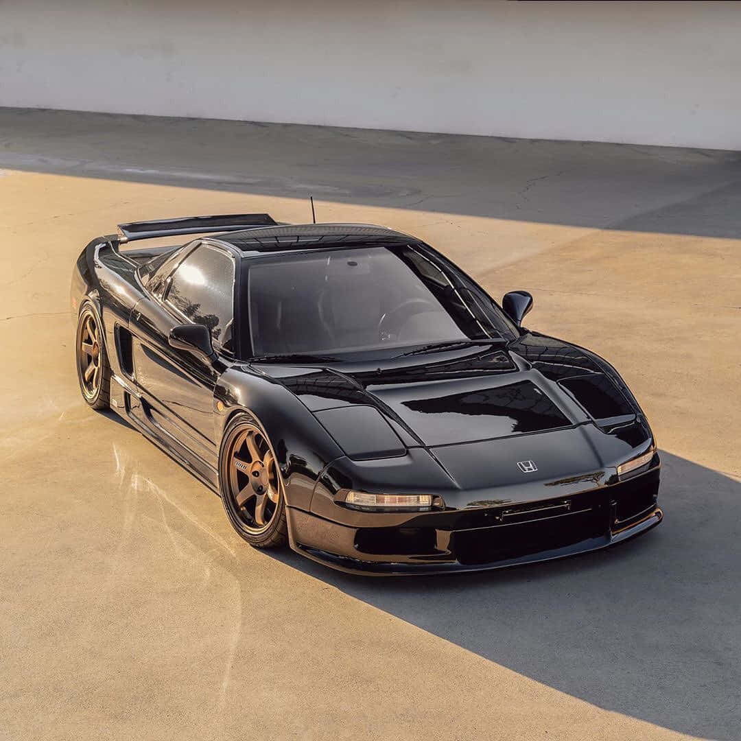 Honda Nsx 桌布 壁紙 — 一輛亮黑色本田NSX跑車配金色輪圈，停泊在陽光普照的混凝土表面上，投下長長的影子。