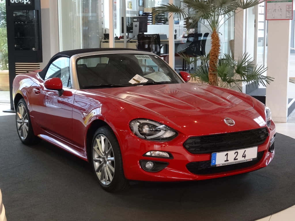 Fiat 124 Spider 桌布 壁紙 — 一輛亮紅色的菲亞特124 Spider開篷車在棕櫚樹旁的展廳燈光下閃閃發光。