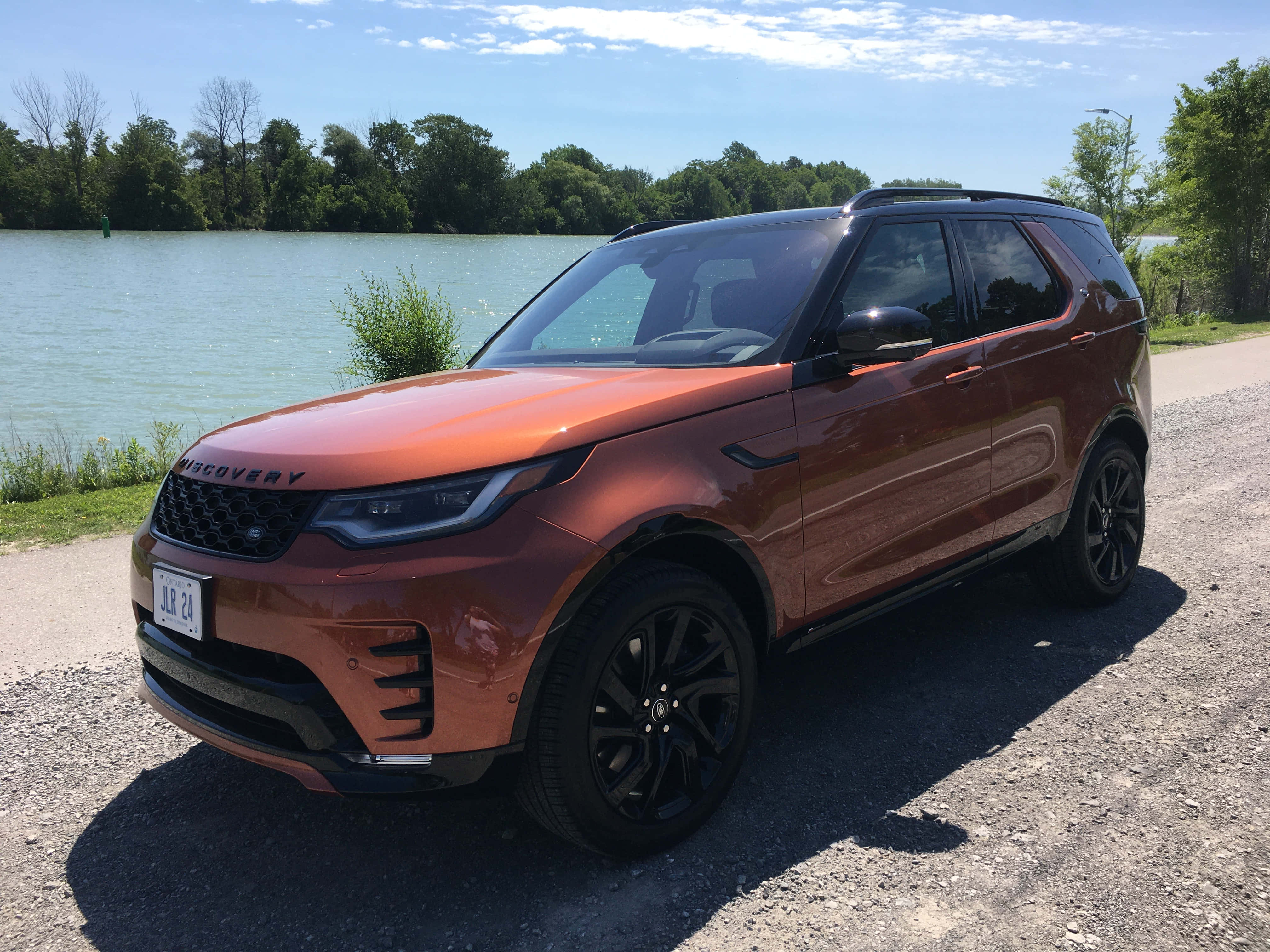 Land Rover Discovery 桌布 壁紙 — 一輛亮面銅色 Land Rover Discovery 停泊在碎石路上，旁邊是平靜的湖泊，上方是晴朗的藍天，點綴著幾朵白雲。