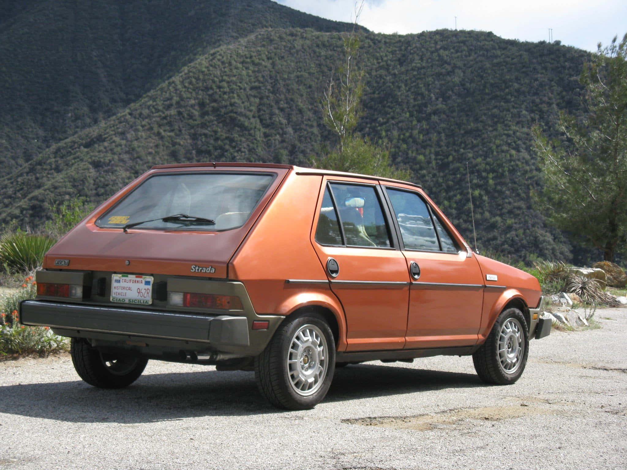 Fiat Ritmo 桌布 壁紙 — 一輛生鏽的橙色 Fiat Strada 掀背車停在碎石路上，背景是明亮的日光下的綠色山丘和樹木。
