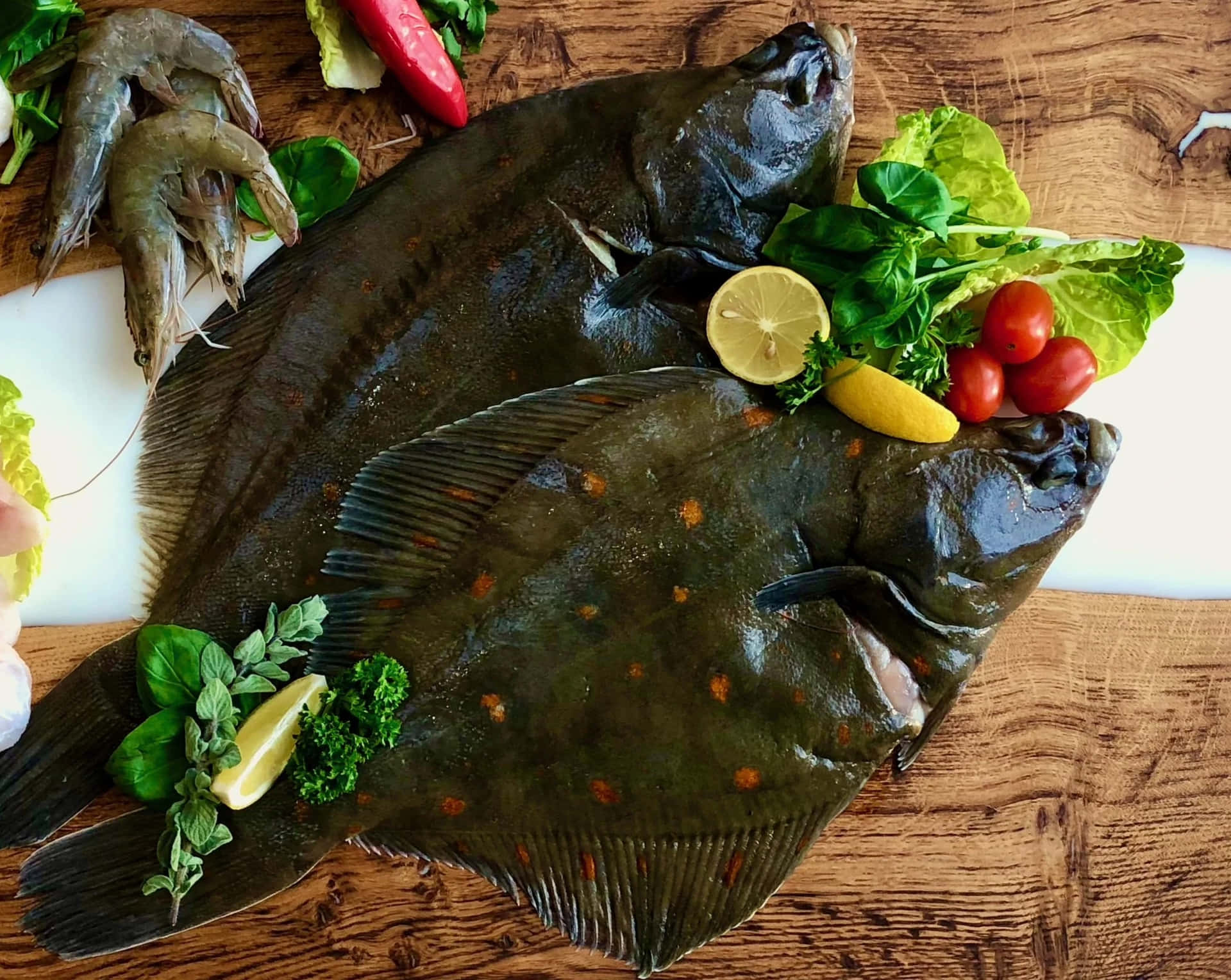  壁紙 — 兩條新鮮的比目魚放在木板上，周圍有蝦、車厘茄、檸檬片和新鮮香草。