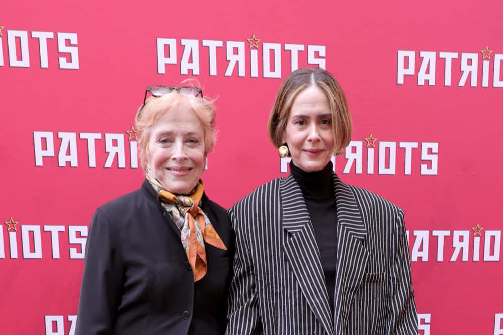  壁紙 — 兩位女士在活動中微笑擺姿勢，背景是紅色的 'PATRIOTS' 品牌背景板。
