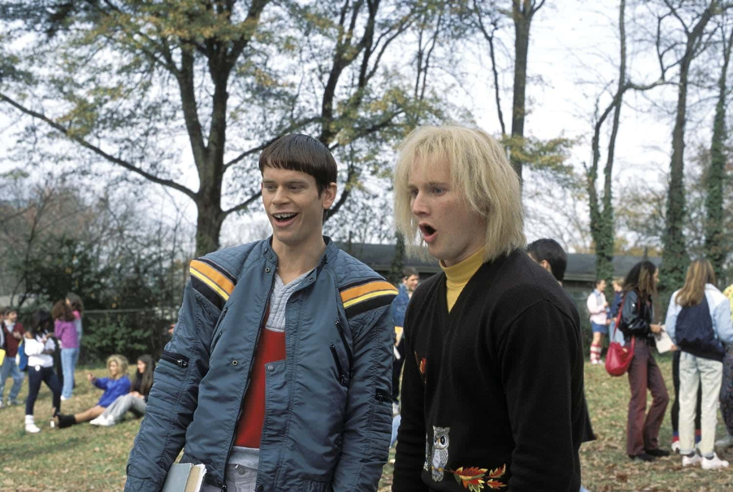  壁紙 — 兩位身穿1980年代時尚的年輕男子站在戶外公園，背景有光禿的樹木和其他人。