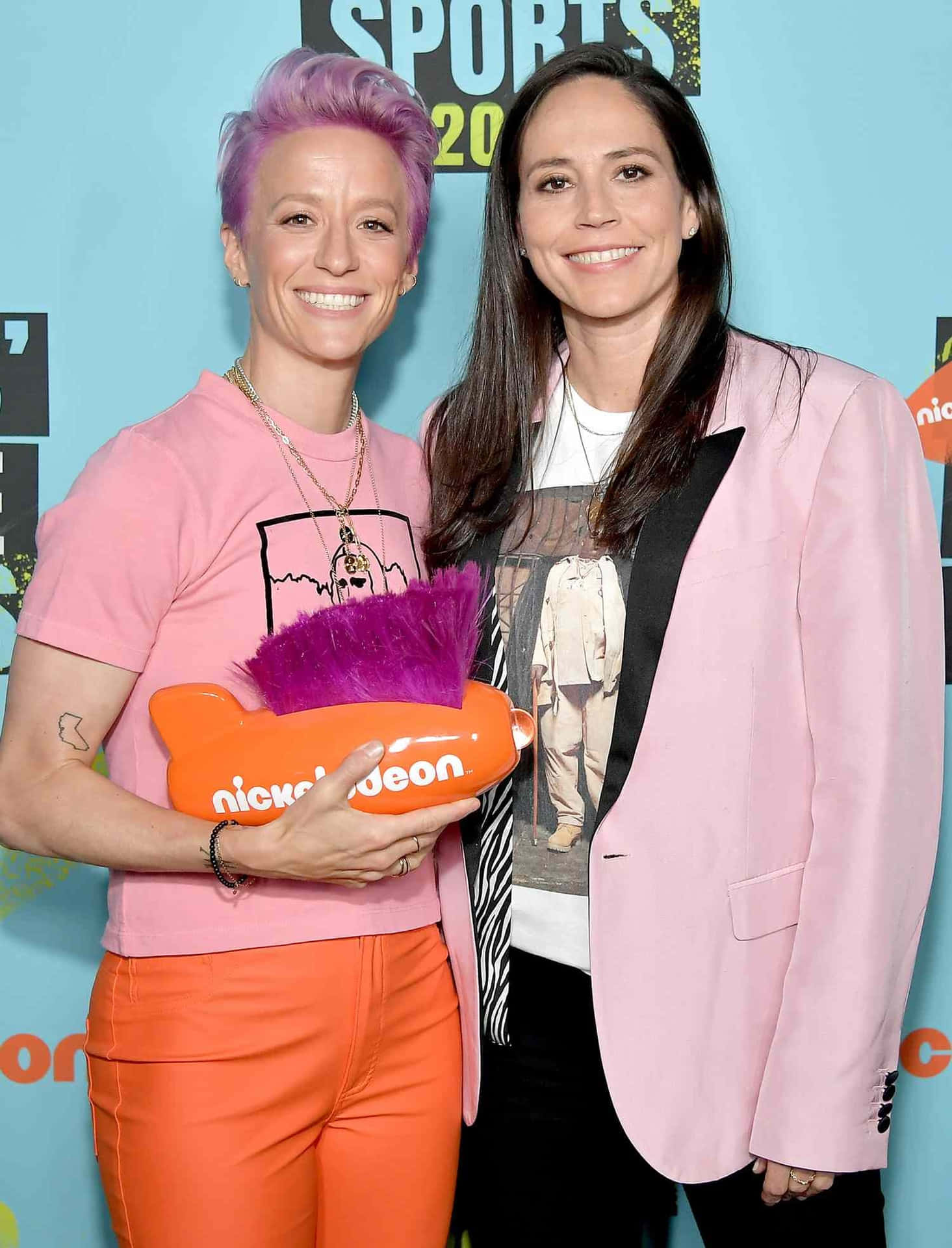 壁紙 — 兩位微笑的女性在Nickelodeon活動中擺姿勢，其中一人手持一個帶有紫色羽毛的亮橙色毛絨獎品。