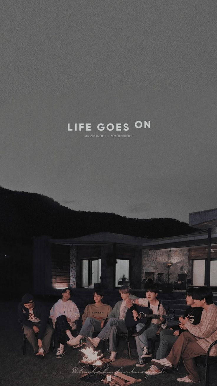 Life Goes On 灰調鎖屏 BTS 壁紙 — 一群年輕男子在黃昏時圍著營火，一人彈結他，背後有山和現代房屋。