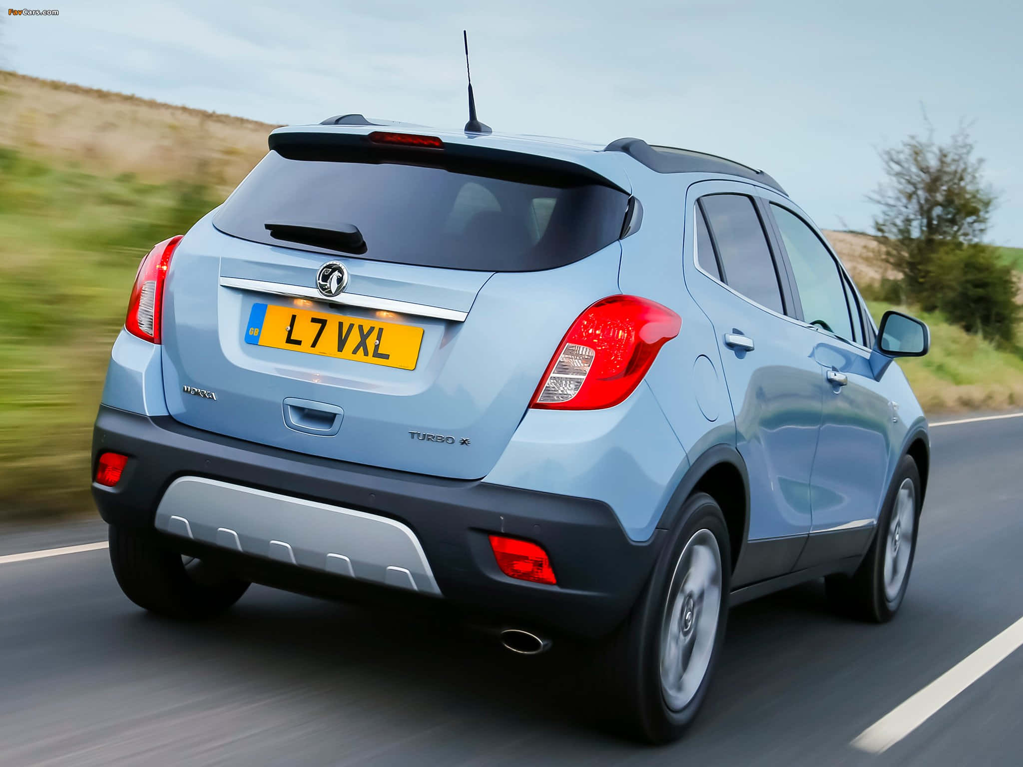 Opel Mokka 桌布 壁紙 — 一輛淺藍色的Vauxhall Mokka Turbo X SUV在晴朗的天空下行駛在鄉村道路上，背景是模糊的綠色山坡。