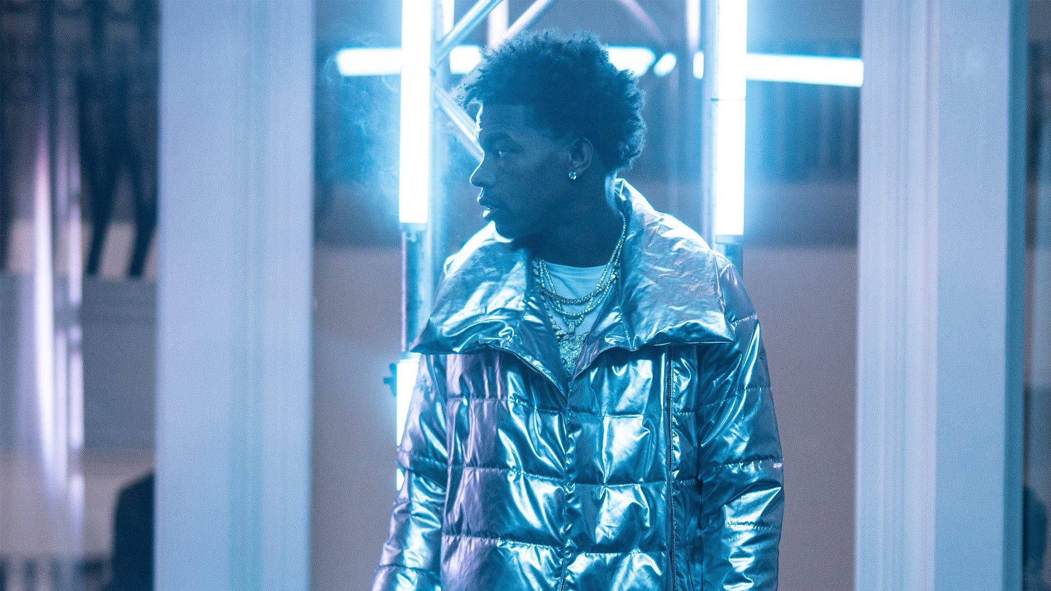 Lil Baby 銀色羽絨外套 壁紙 — 一位身穿閃亮金屬外套的年輕男子站在一條充滿未來感的藍色走廊中，周圍有發光的霓虹燈管。