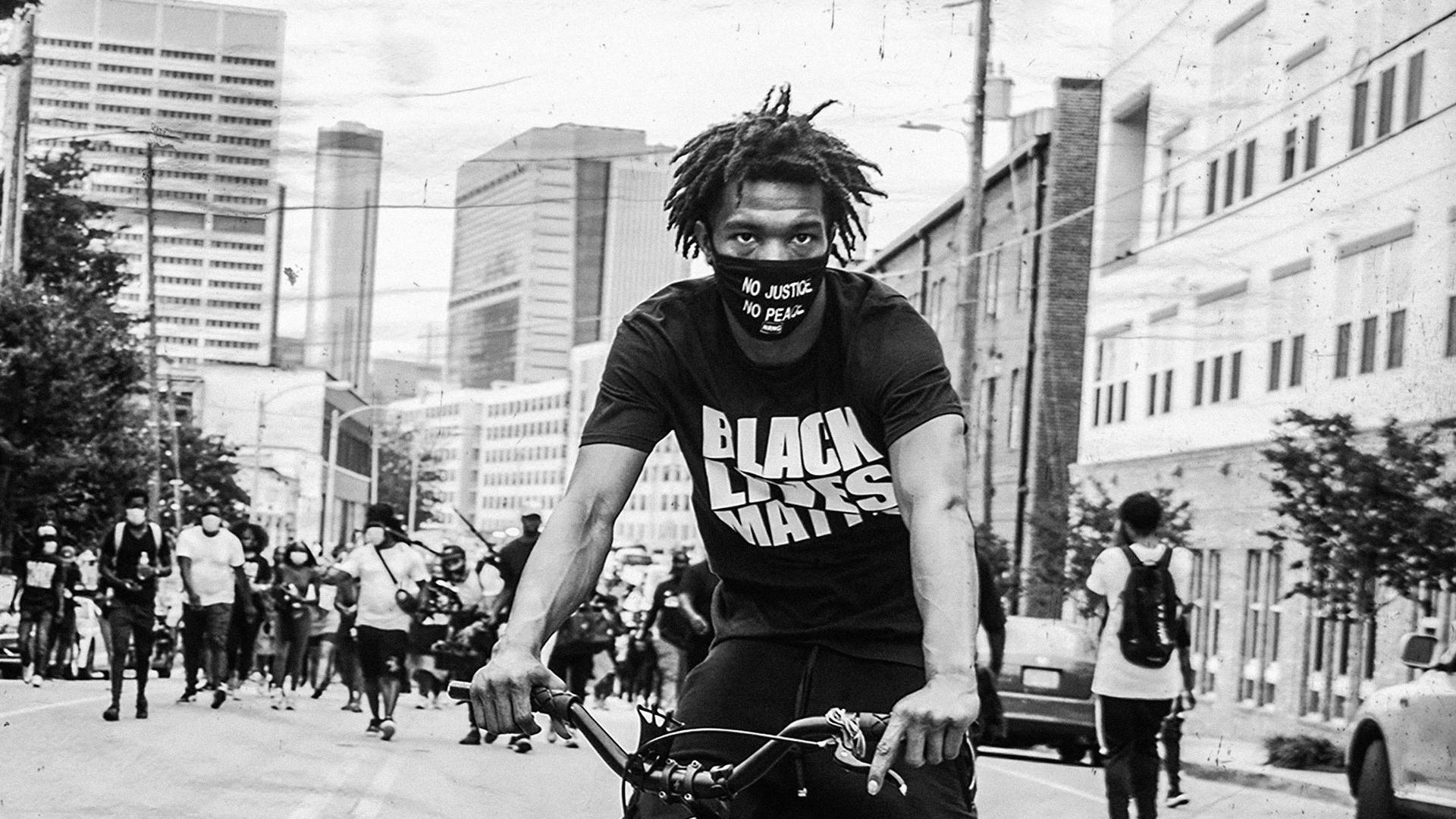 踩單車的黑人男子身穿「Black Lives Matter」T恤和口罩，帶領抗議遊行穿過城市街道。 — 細路仔 GIF 壁紙