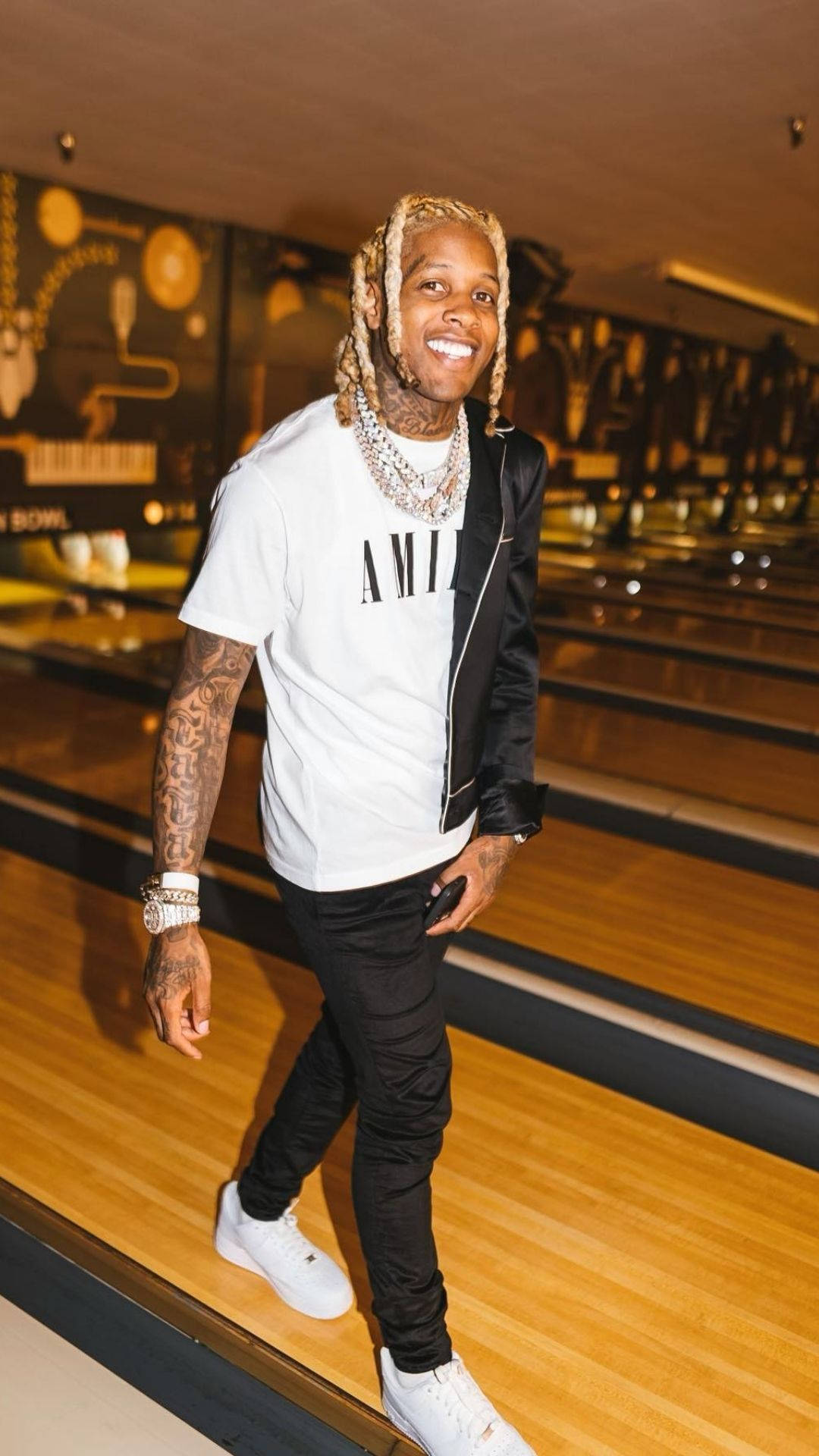 Lil Durk 保齡球館 壁紙 — 一位微笑的男子，梳著辮子頭，身上有紋身，自信地走在拋光的木製保齡球道上，身穿白色T恤、黑色外套和白色運動鞋。