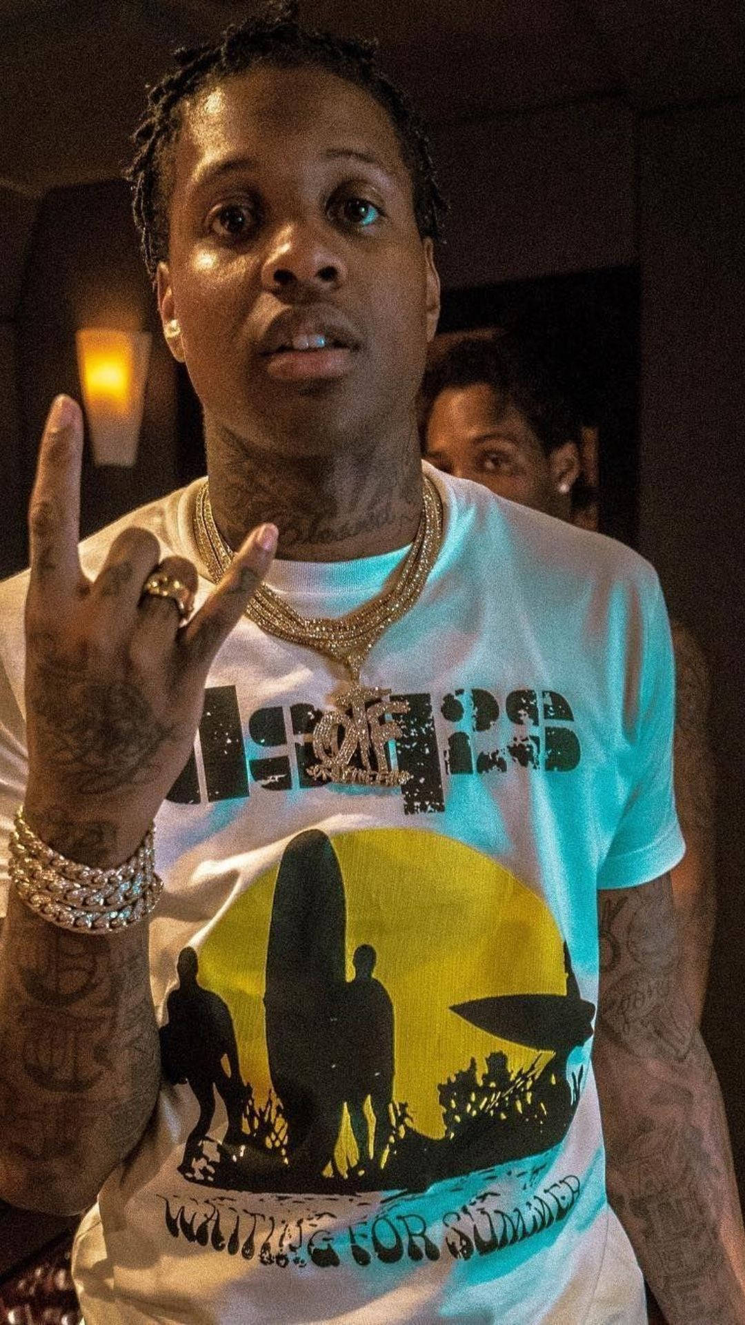 Lil Durk 做拉丁王國饒舌手勢 壁紙 — 一名有紋身和黃金首飾的男子在昏暗的房間裡穿著圖案T恤做手勢。