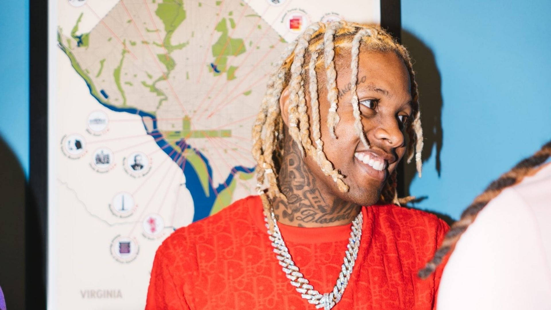 Lil Durk 側面視角 壁紙 — 一位微笑的男子，編著辮子頭髮，有紋身，穿著紅色襯衫和鏈條，背景是維珍尼亞州地圖。