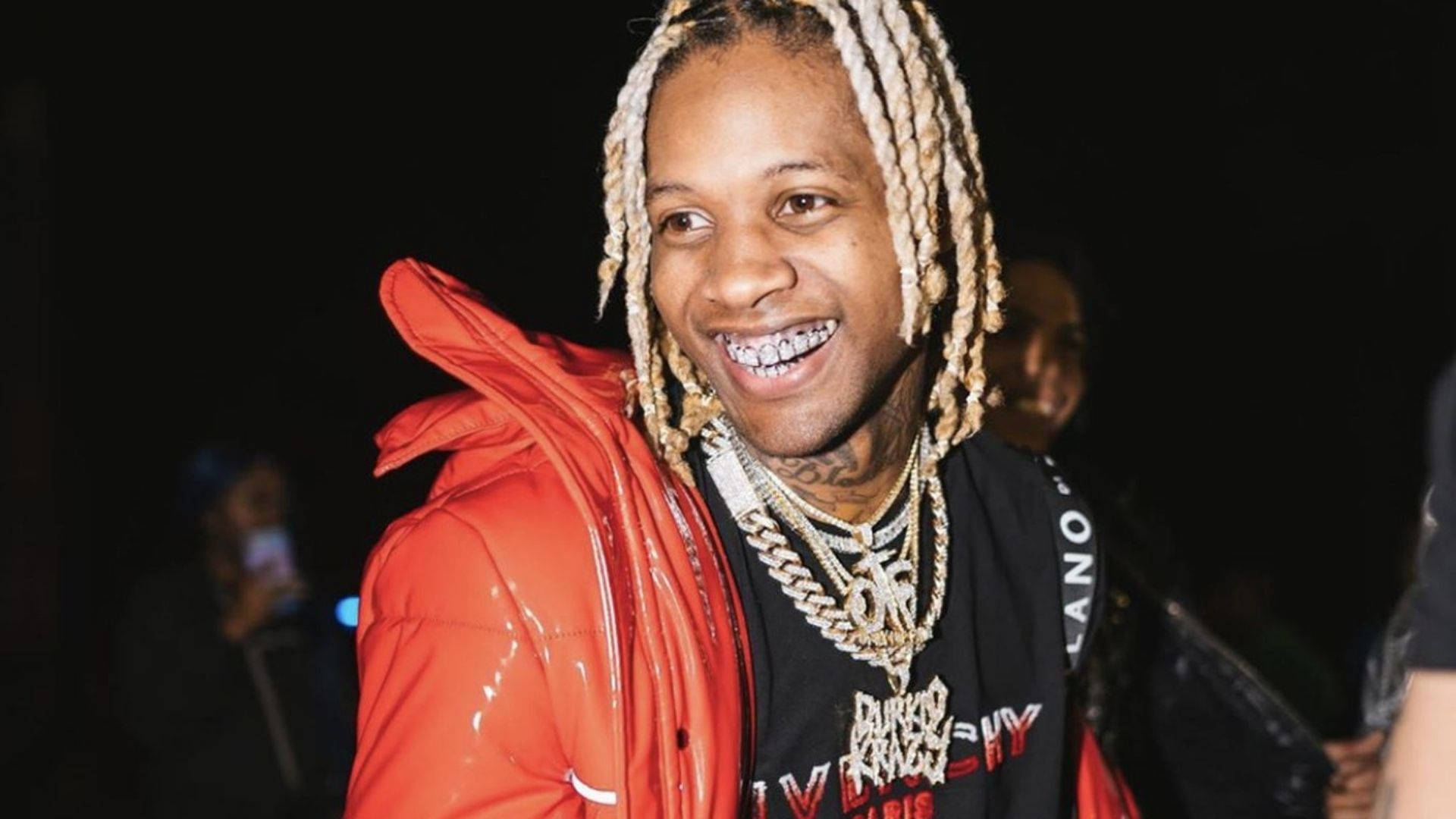 Lil Durk 在人群中微笑 壁紙 — 一位微笑的男子，編著辮子、戴著牙套，身穿亮紅色羽絨外套，層疊金鍊，背景為深色。