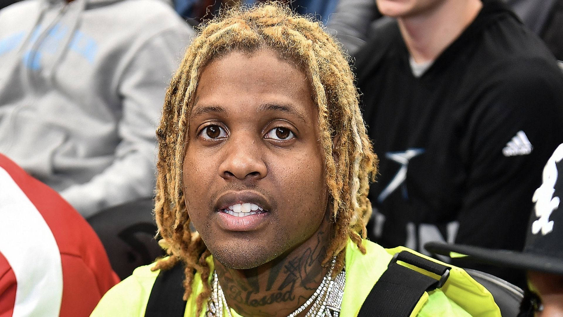 Lil Durk 坐看籃球賽 壁紙 — 一個金髮辮和紋身的男子在擁擠的活動中穿著亮黃色外套的特寫。