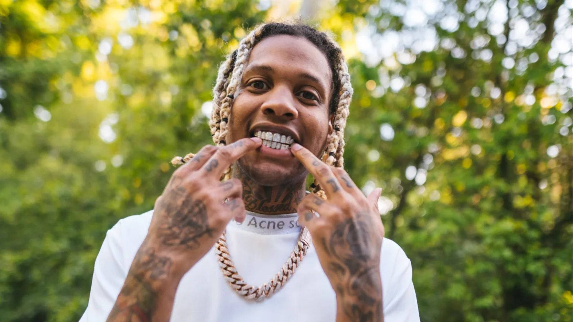 Lil Durk 展露白牙 壁紙 — 一個微笑的男人，梳著辮子頭和紋身，在陽光普照的綠色森林背景下，頑皮地指著他的金牙套。