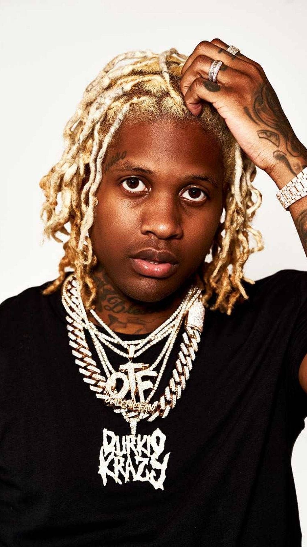 Lil Durk 搭配時尚金髮造型 壁紙 — 一位年輕男子，金色辮子頭和紋身，自信地擺姿勢，戴著多層鑽石鏈條和黑色圖案T恤，背景是純白色。