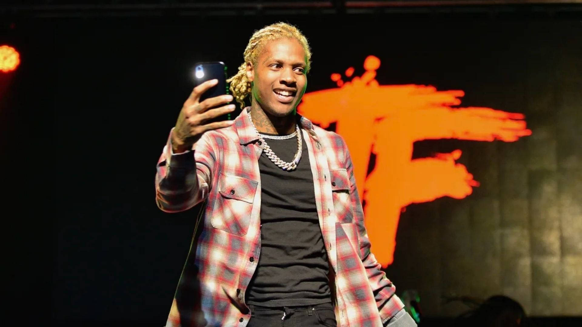 Lil Durk 攝影 壁紙 — 一位微笑的金髮辮子男在明亮的橙色燈光下於舞台上自拍。