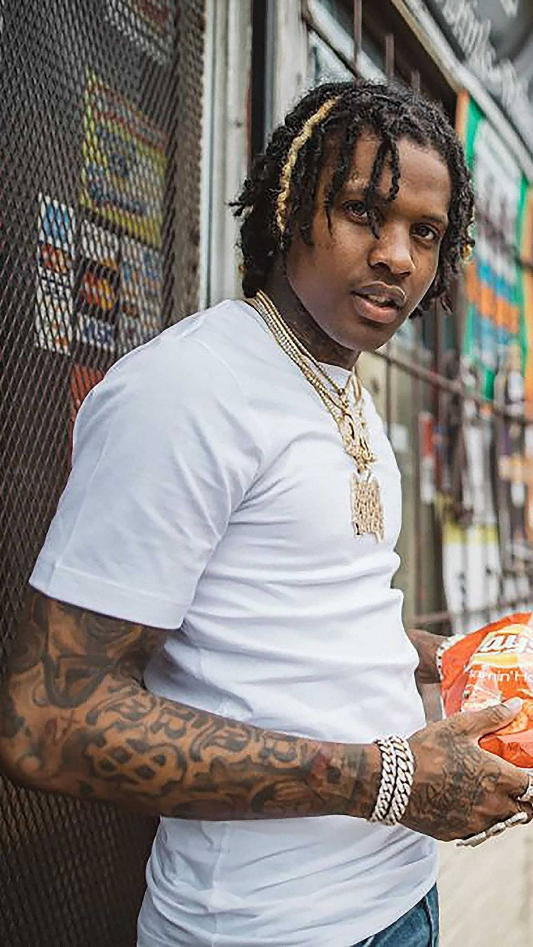 Lil Durk 模特造型 壁紙 — 一位辮髮、有紋身的年輕男子在都市小巷中靠著鐵絲網圍欄，手持一包薯片。