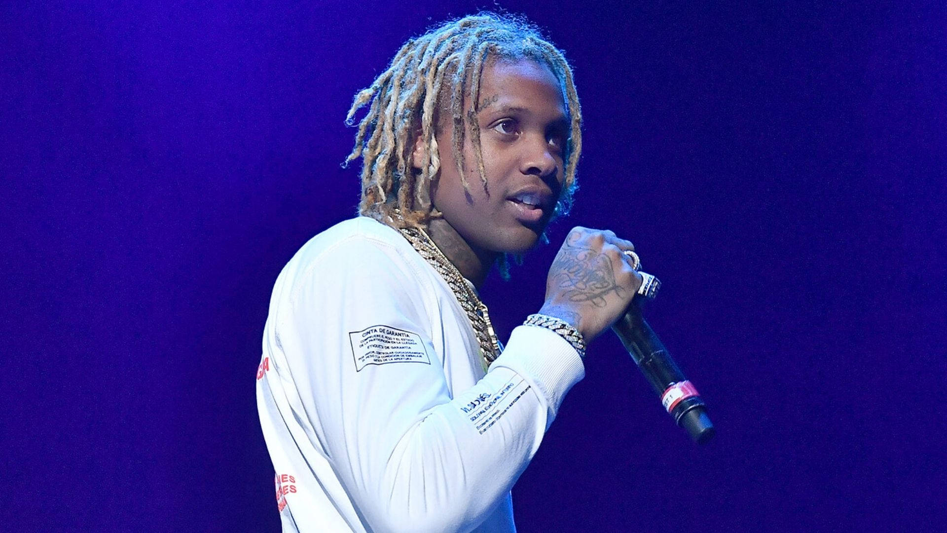 Lil Durk 演唱中 壁紙 — 一位梳著辮子的說唱歌手在紫色燈光下拿著麥克風在舞台上表演。