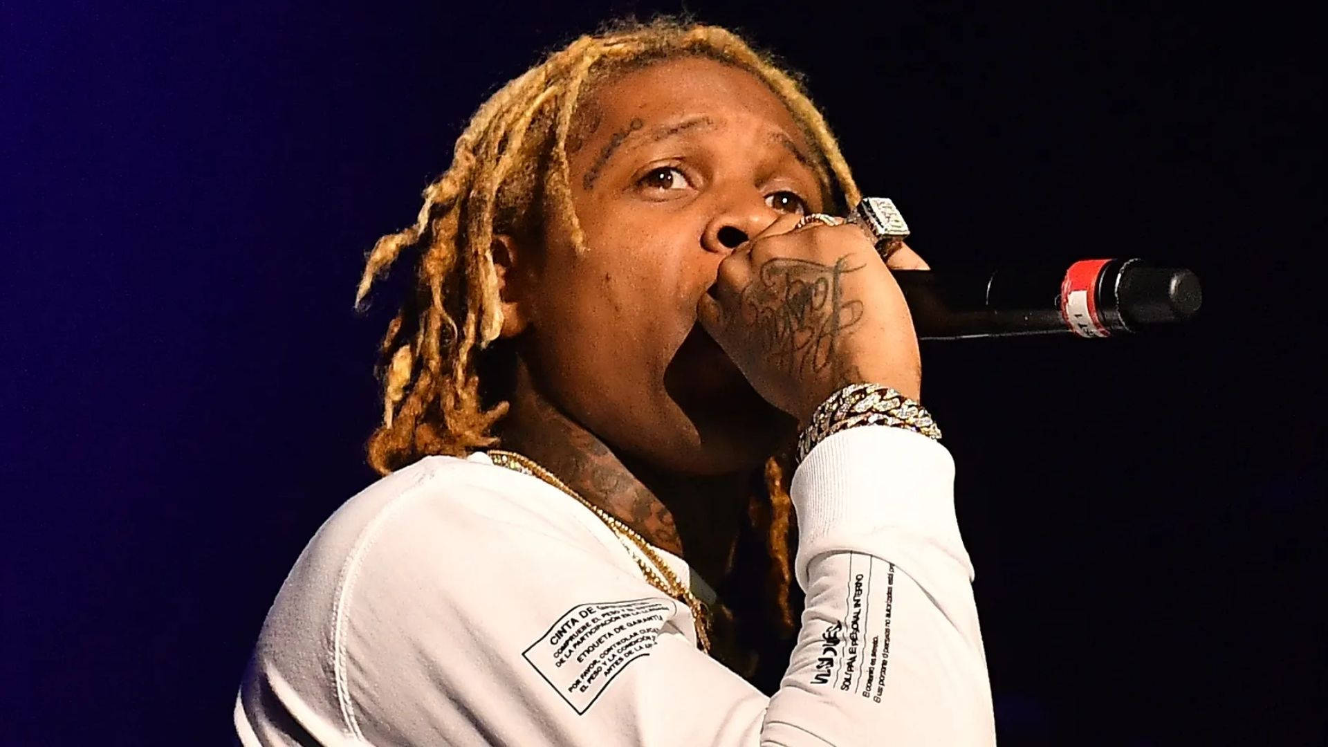 Lil Durk 演唱會熱情演出瞬間 壁紙 — 金髮辮子說唱歌手在藍色舞台燈光下，穿著白襯衫和戴金鏈，對著麥克風唱歌。