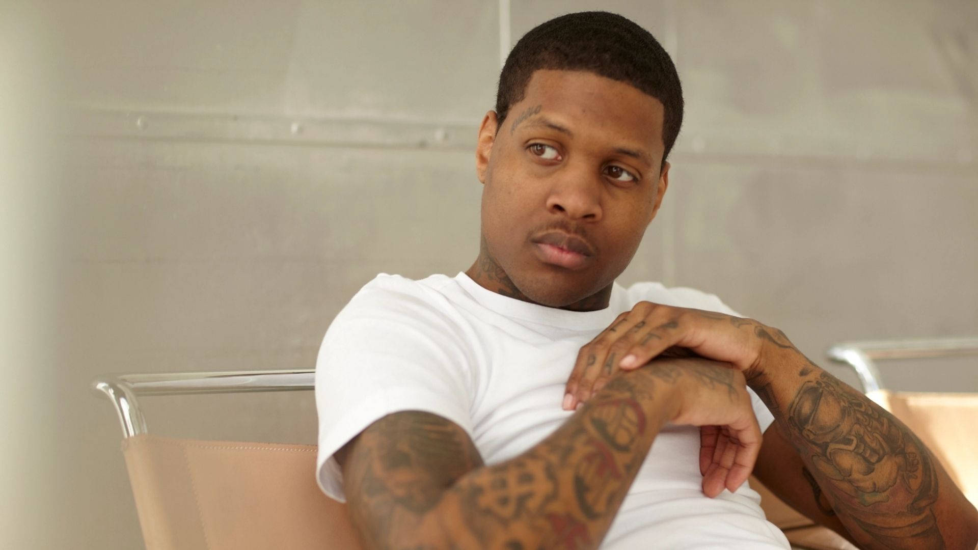 Lil Durk 白色T恤造型 壁紙 — 一個有紋身手臂的男人坐在椅子上，穿著白色T恤，嚴肅地望向一邊。