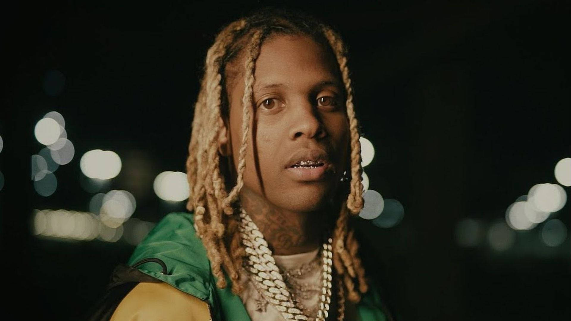 Lil Durk 背景散景燈光 壁紙 — 一個辮子頭髮、戴著金鏈的男人，在模糊的夜間城市景觀和發光的散景燈光前專注地凝視。